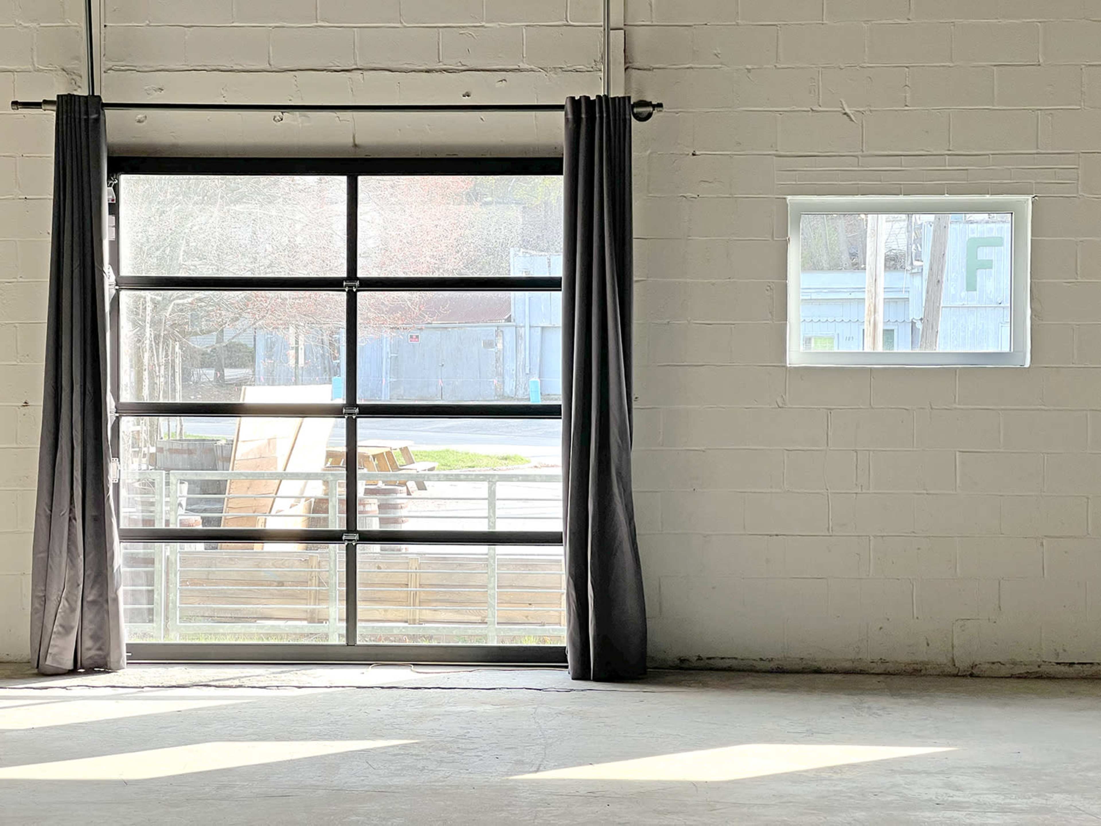 An industrial space with a large glass door and a smaller window on a white, concrete wall, both covered by dark curtains.