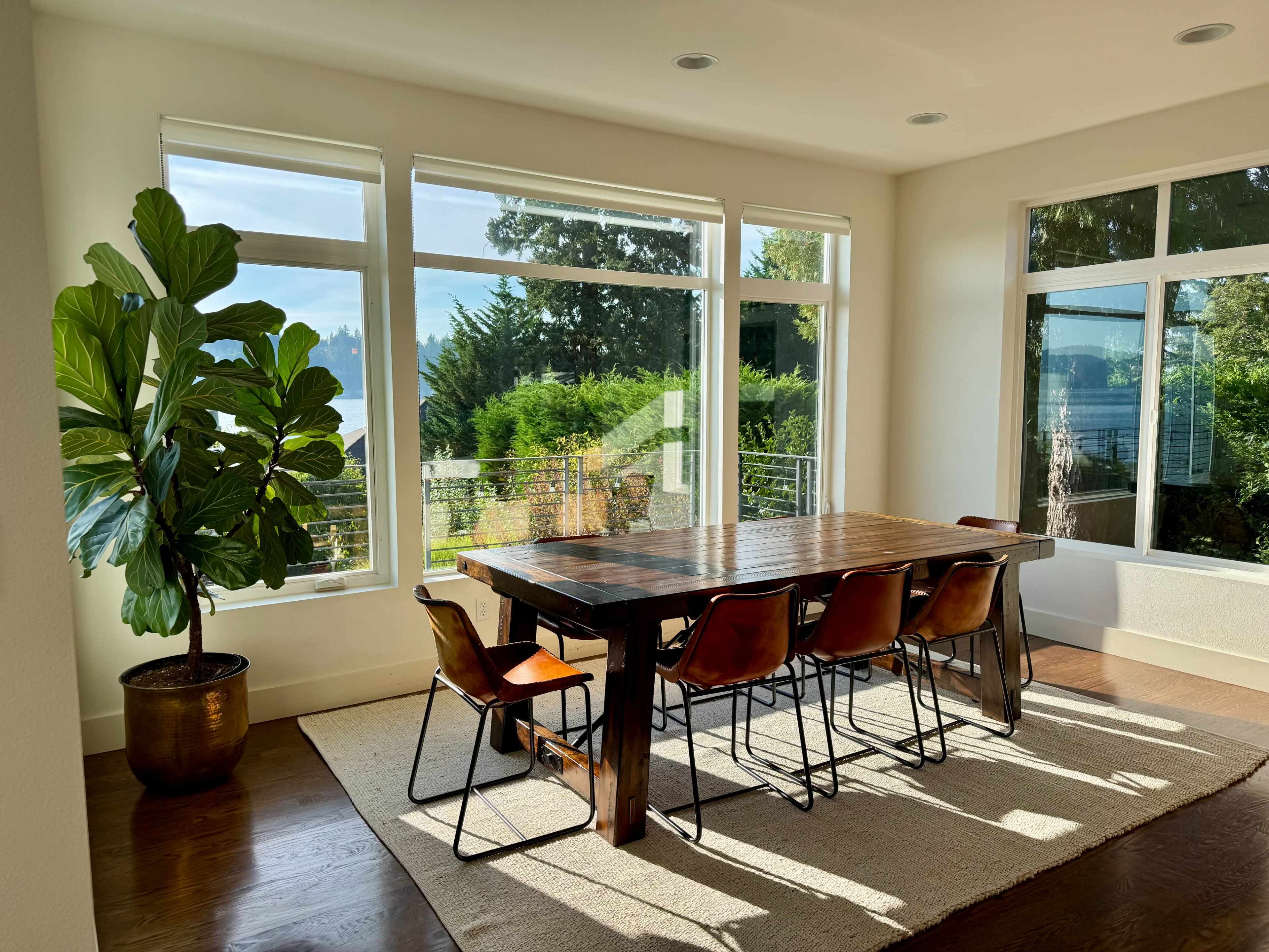 A wooden dining table surrounded by chairs is set in a bright room with large windows overlooking a garden.