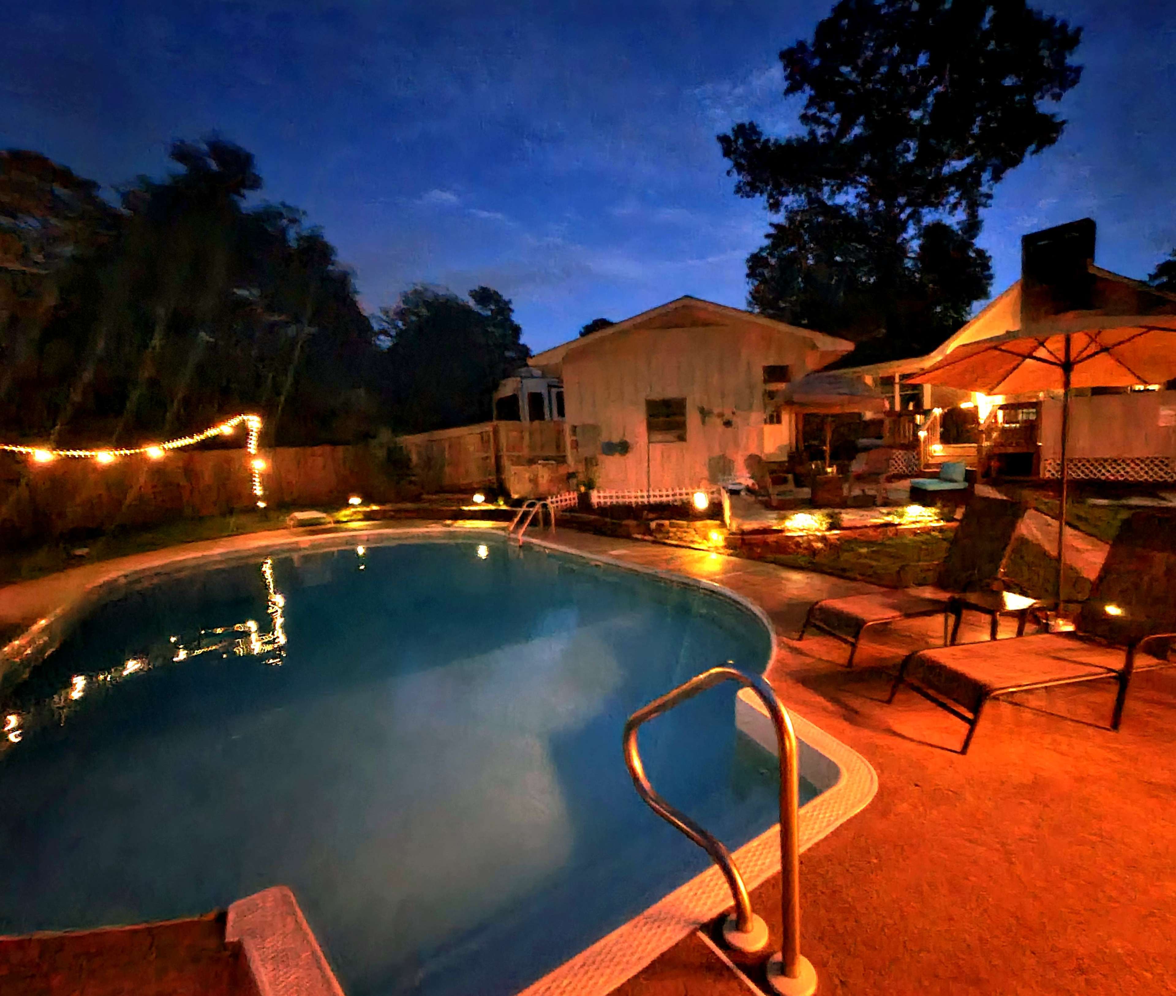 A pool area is illuminated by lights at night, surrounded by lounge chairs and a house in the background.