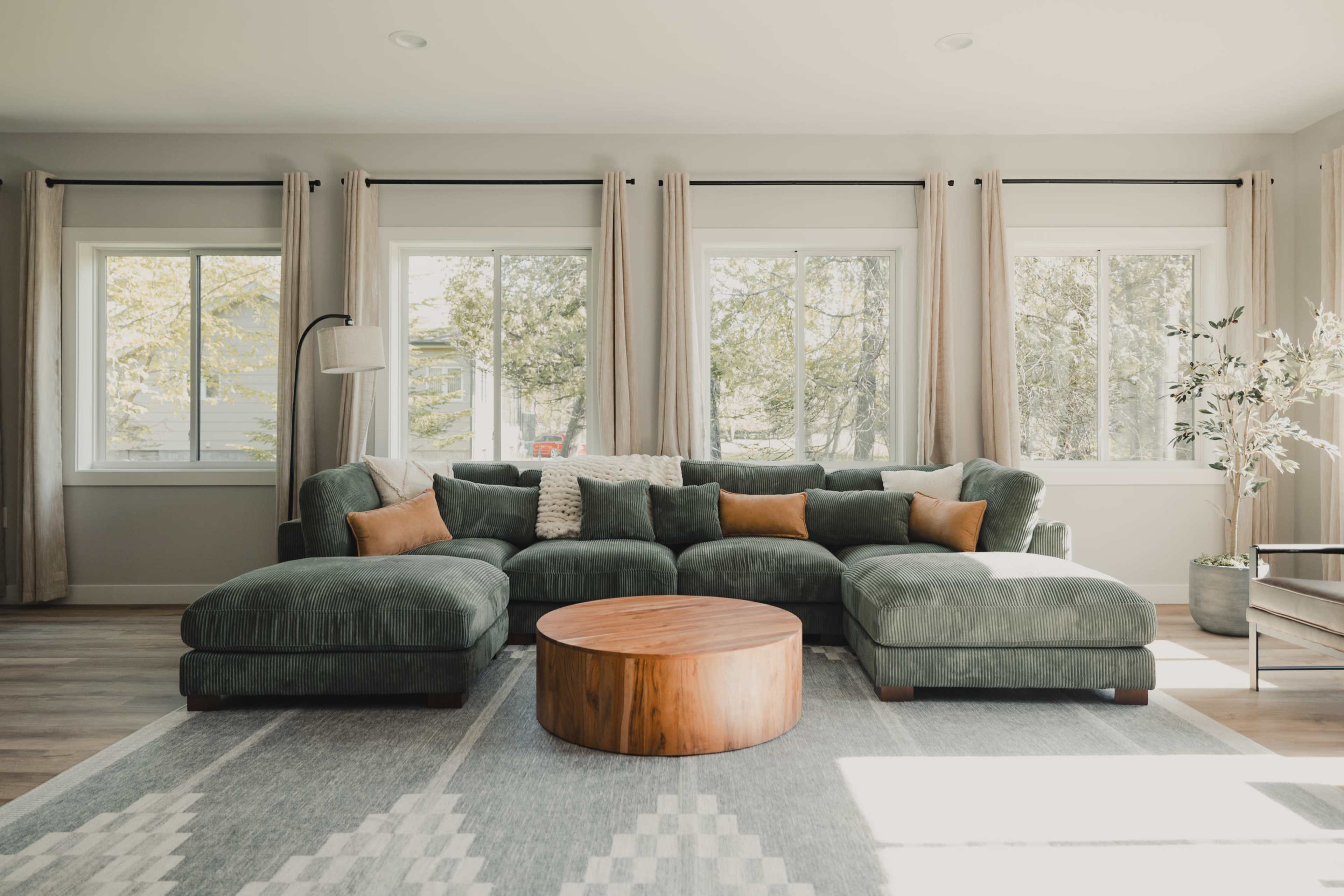 A green sectional sofa with brown accent pillows is placed in a brightly lit living room with large windows and a round wooden coffee table.