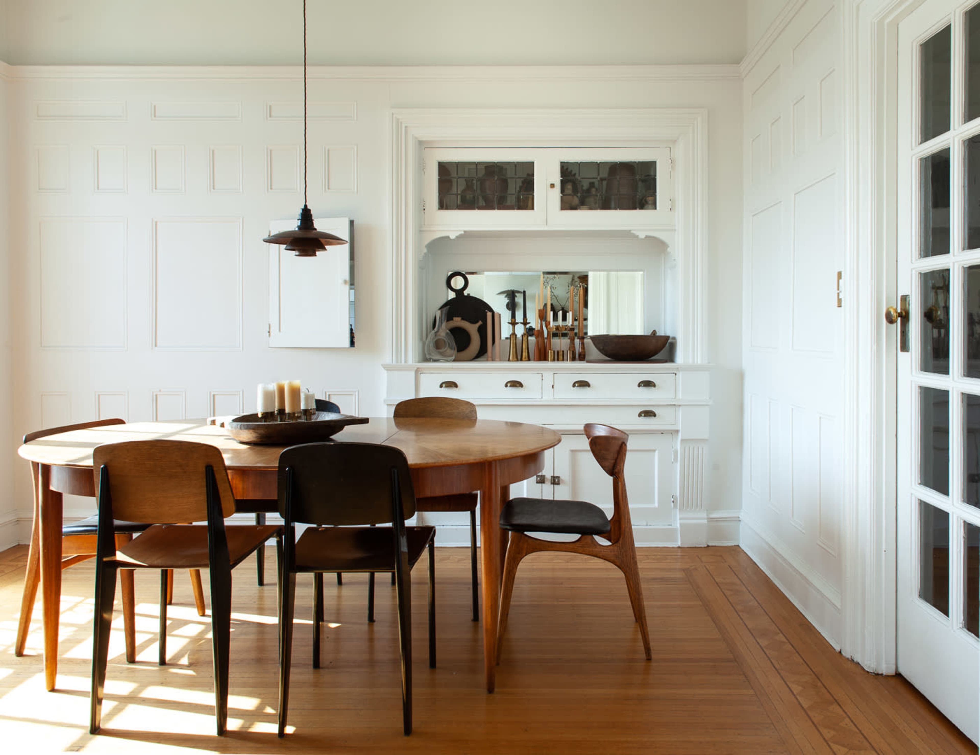 A round wooden dining table with four chairs is positioned in a bright room with white walls and a large cabinet against one side.