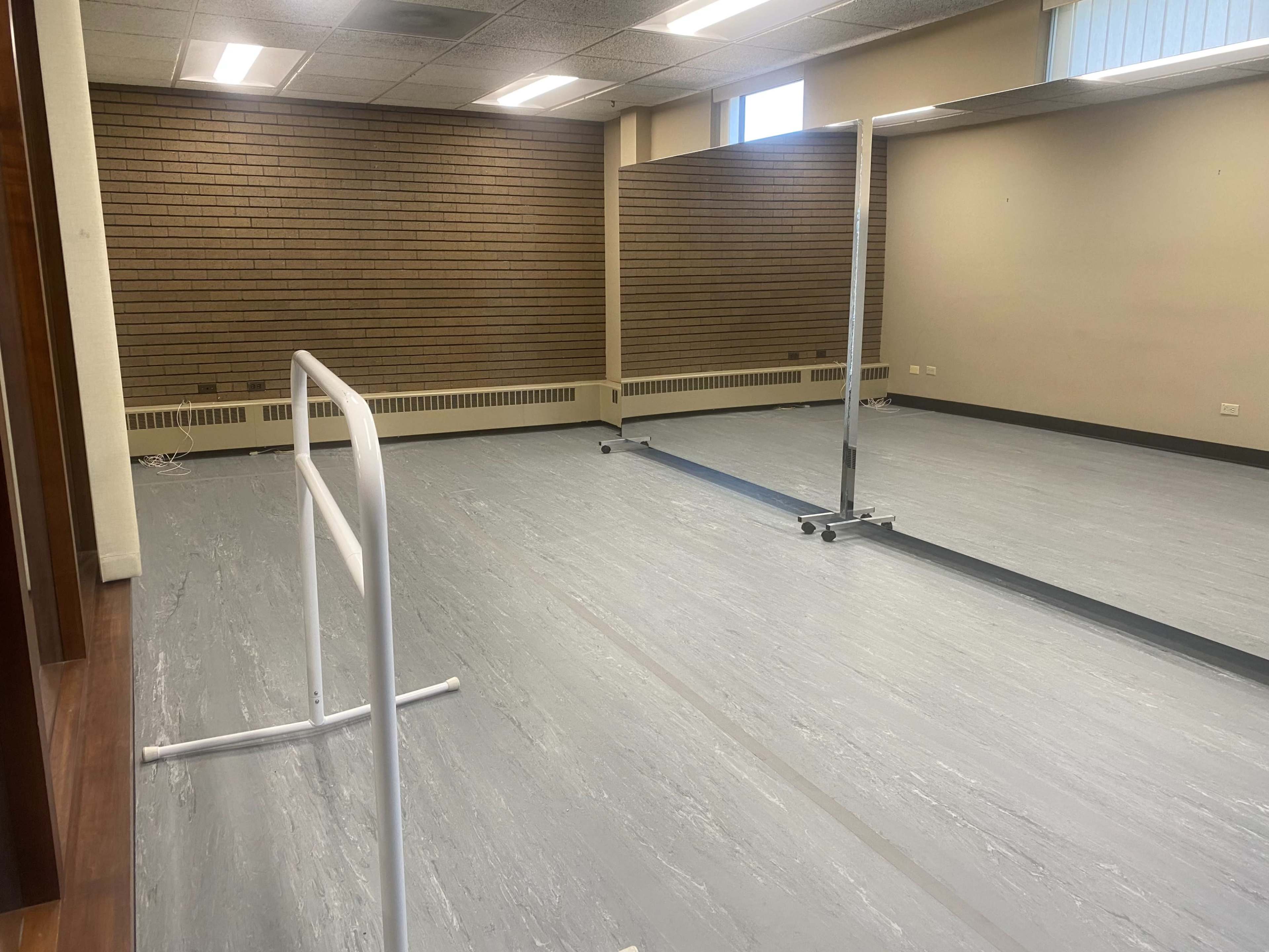 A spacious dance studio with a mirror, ballet barre, and grey flooring.