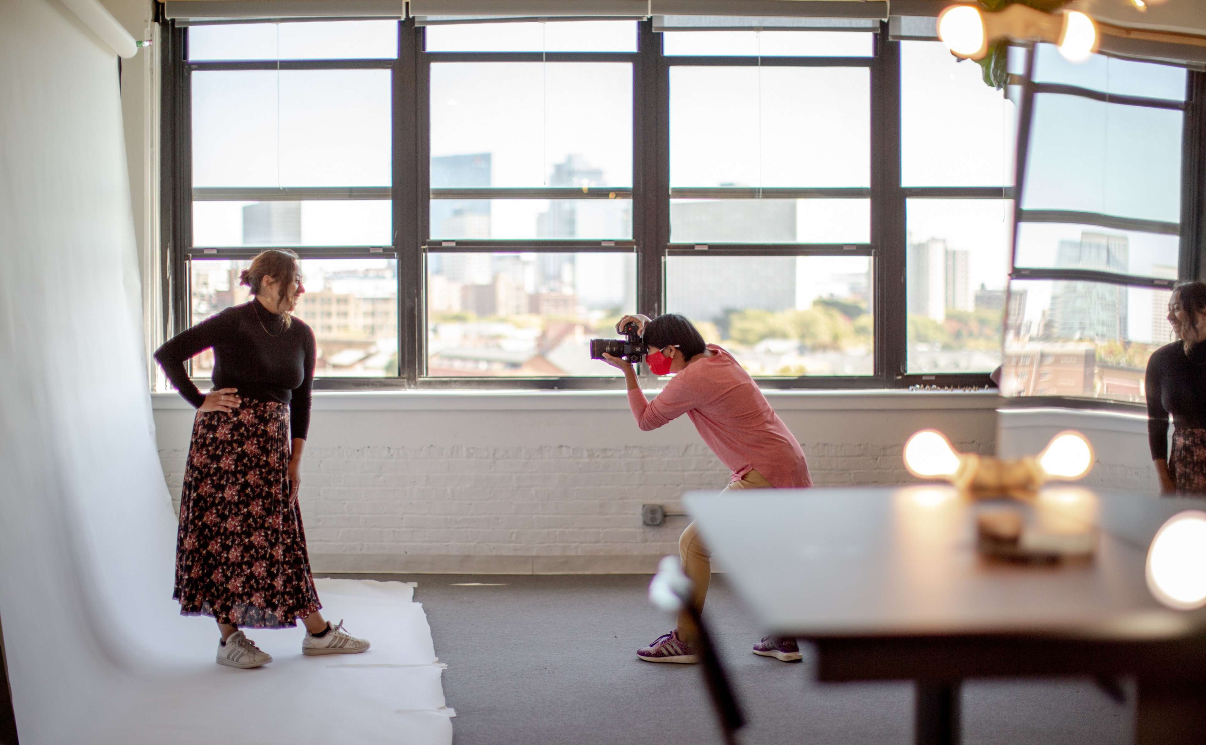 A photographer captures a model posing with a floral skirt in a sunlit studio with large windows and a city skyline view.
