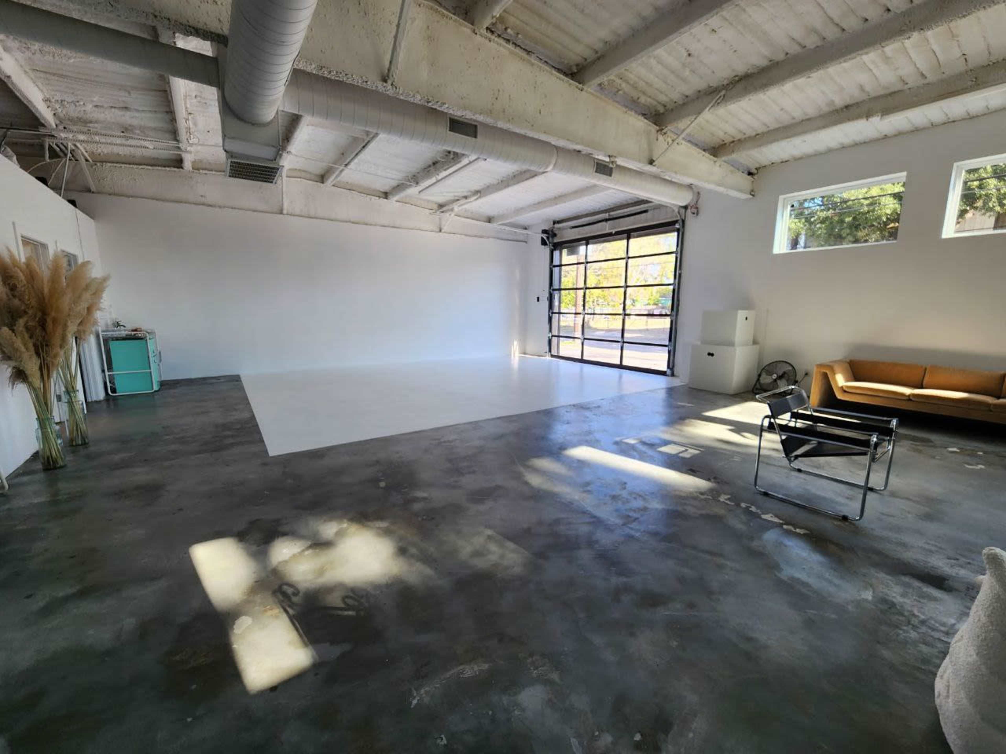 A spacious, empty studio with a polished concrete floor, large windows, and minimal furniture, including a couch and a chair.