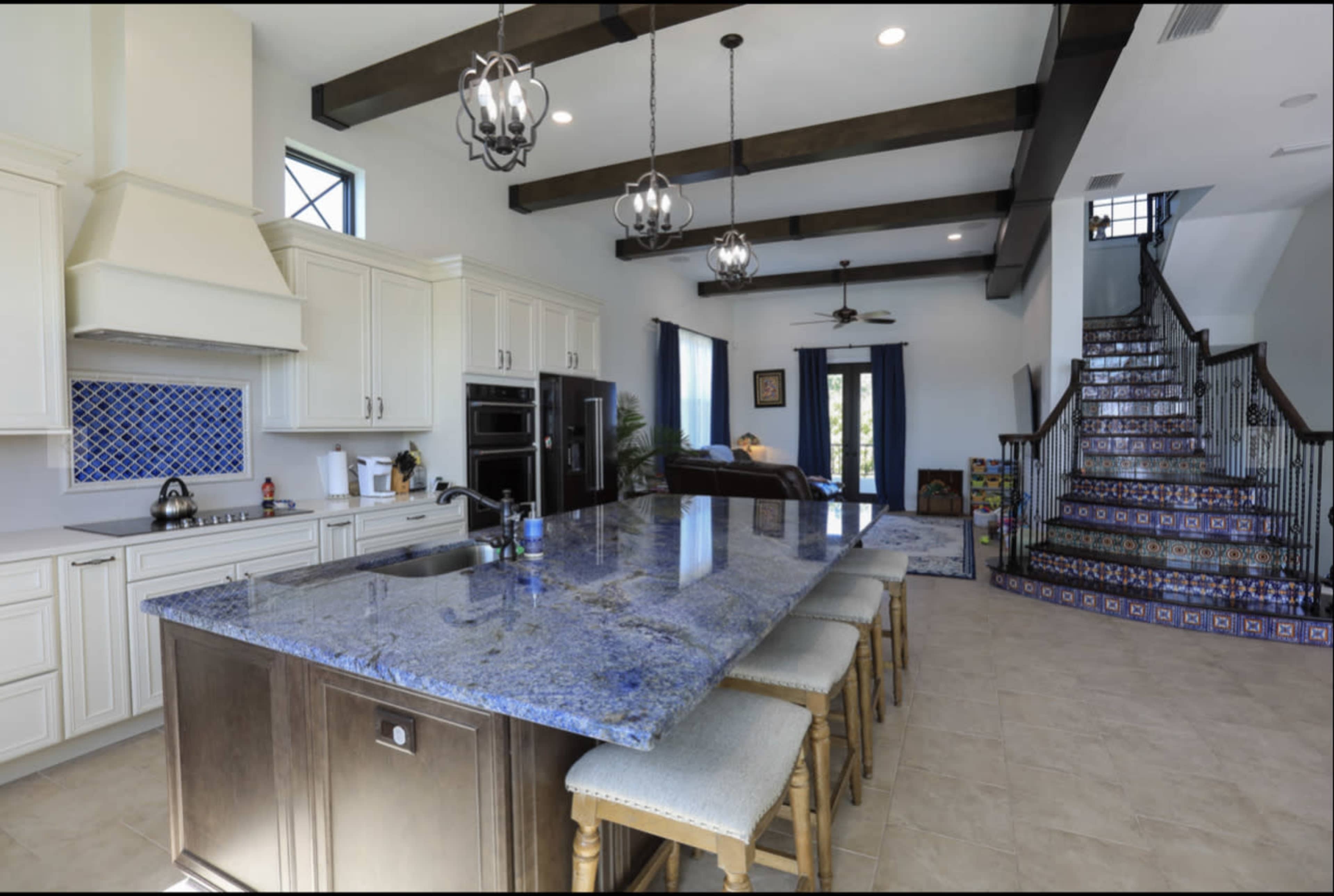 The image shows a modern kitchen with a large island, upper cabinets, and a staircase leading to a second floor, featuring decorative tiles.