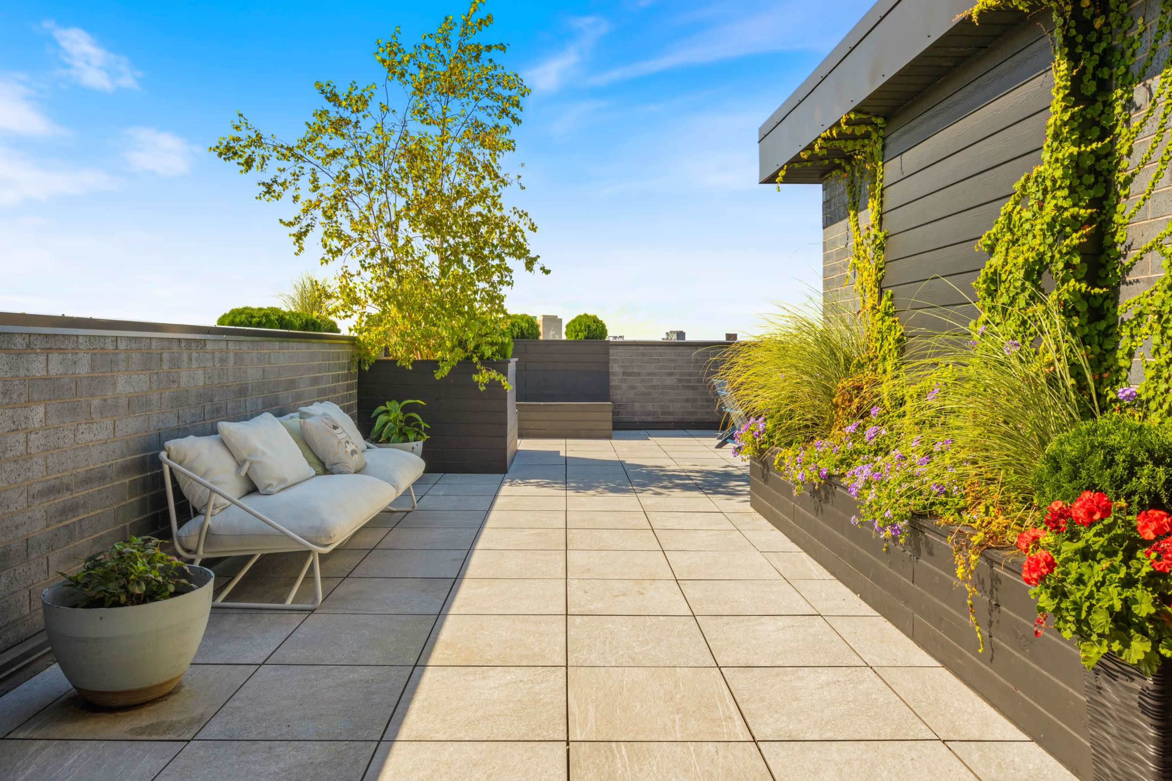 The image shows a rooftop terrace with gray stone tiles, potted plants, a green wall, and a seating area featuring white cushions.