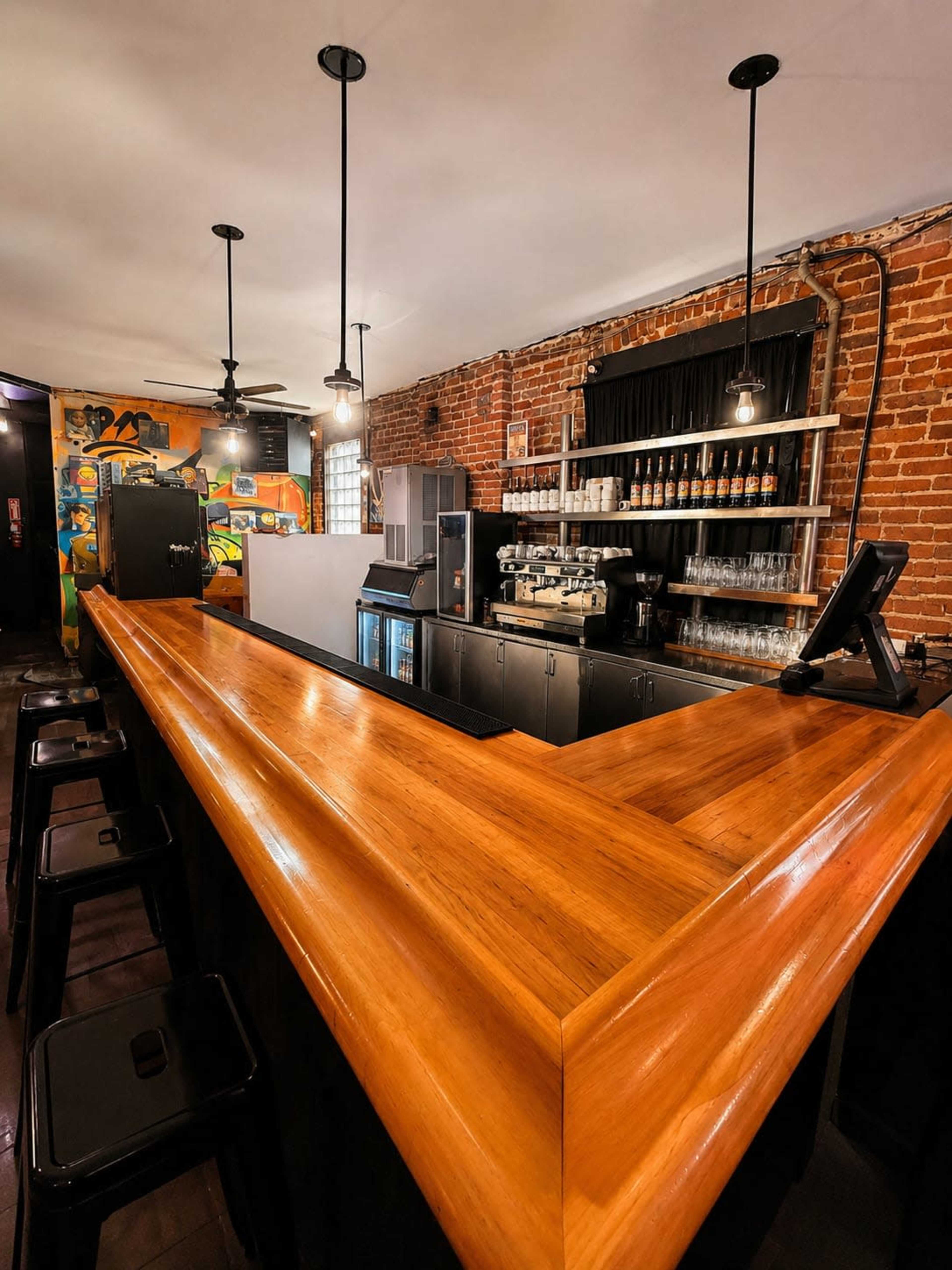 The image shows a wooden bar with high stools, situated in a brick-walled interior that features a coffee machine and glassware on display.