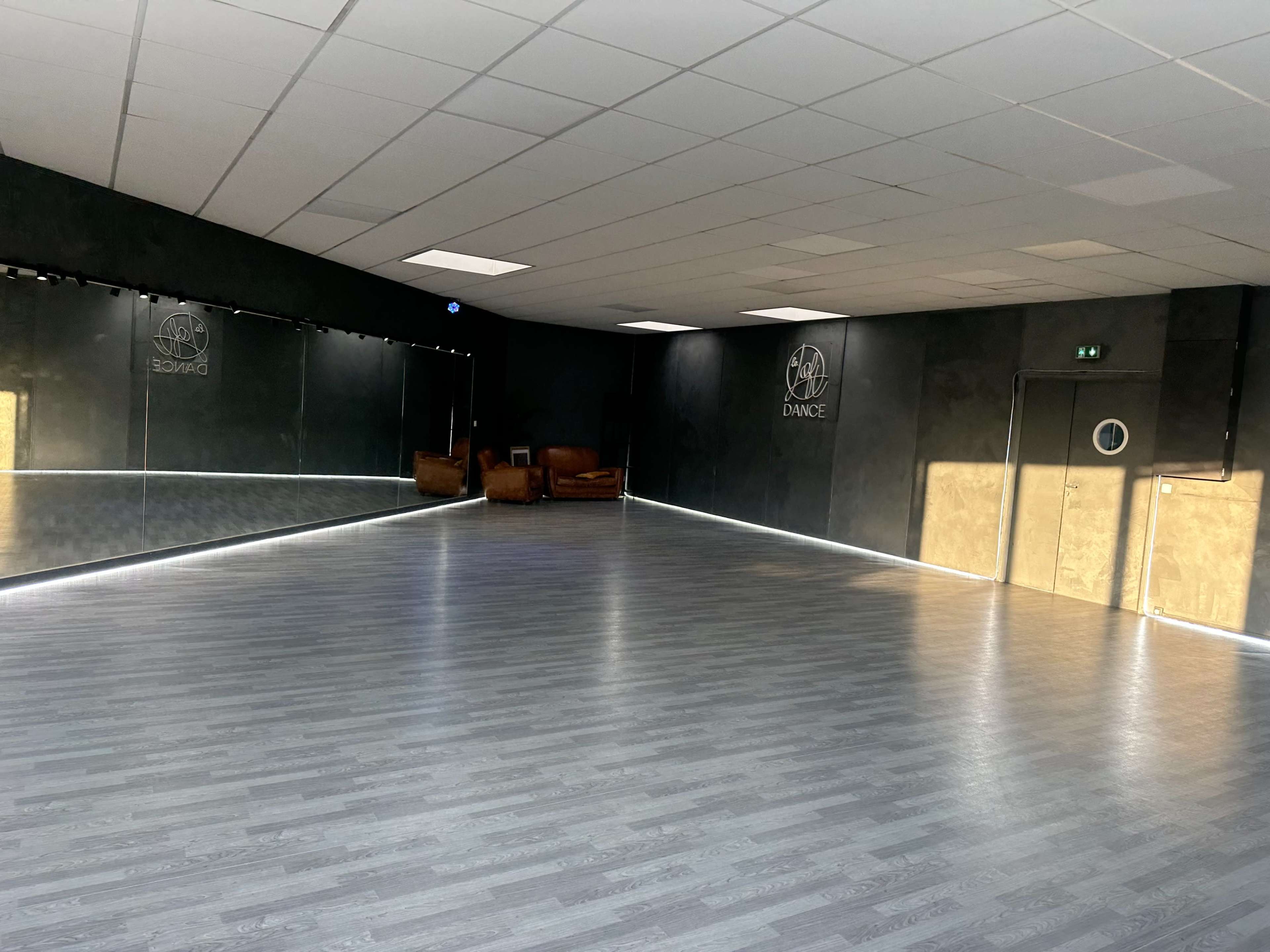 The image features a spacious dance studio with a large mirrored wall, wooden flooring, and a couple of brown chairs in the corner.