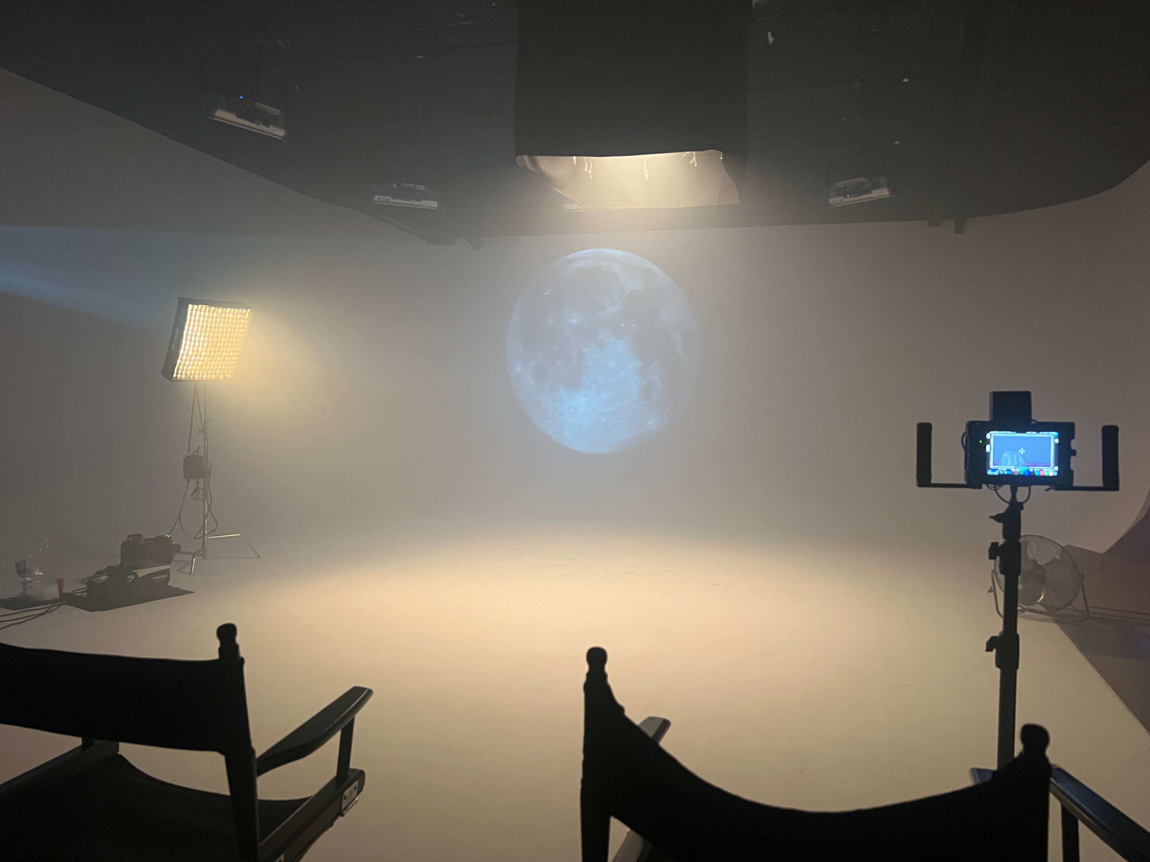 The image shows a dimly lit studio space with a large projection of the moon on a plain background, accompanied by studio lights and two empty chairs.