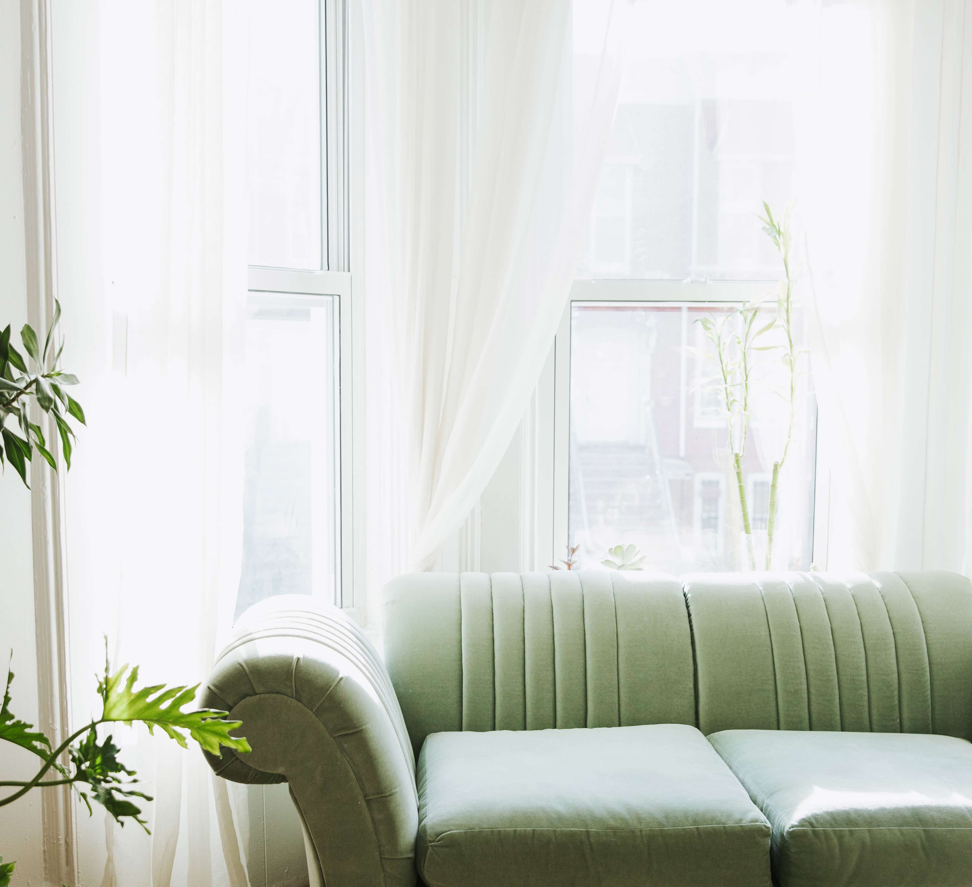 A green couch sits beside a large window adorned with sheer white curtains, allowing natural light to fill the room.