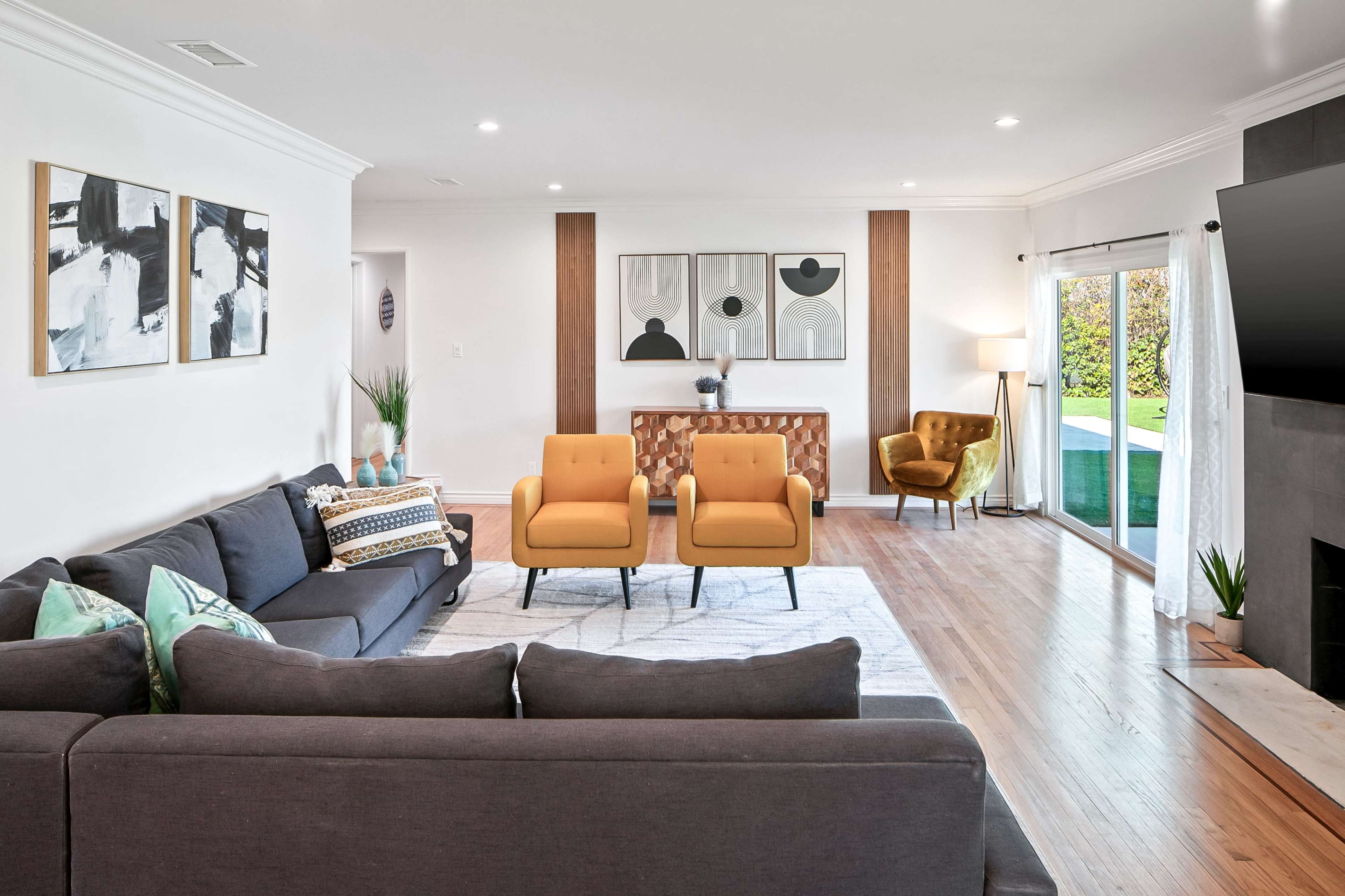 The living room features a gray sectional sofa, two yellow armchairs, and a console table against the wall, with large windows providing a view of the garden.