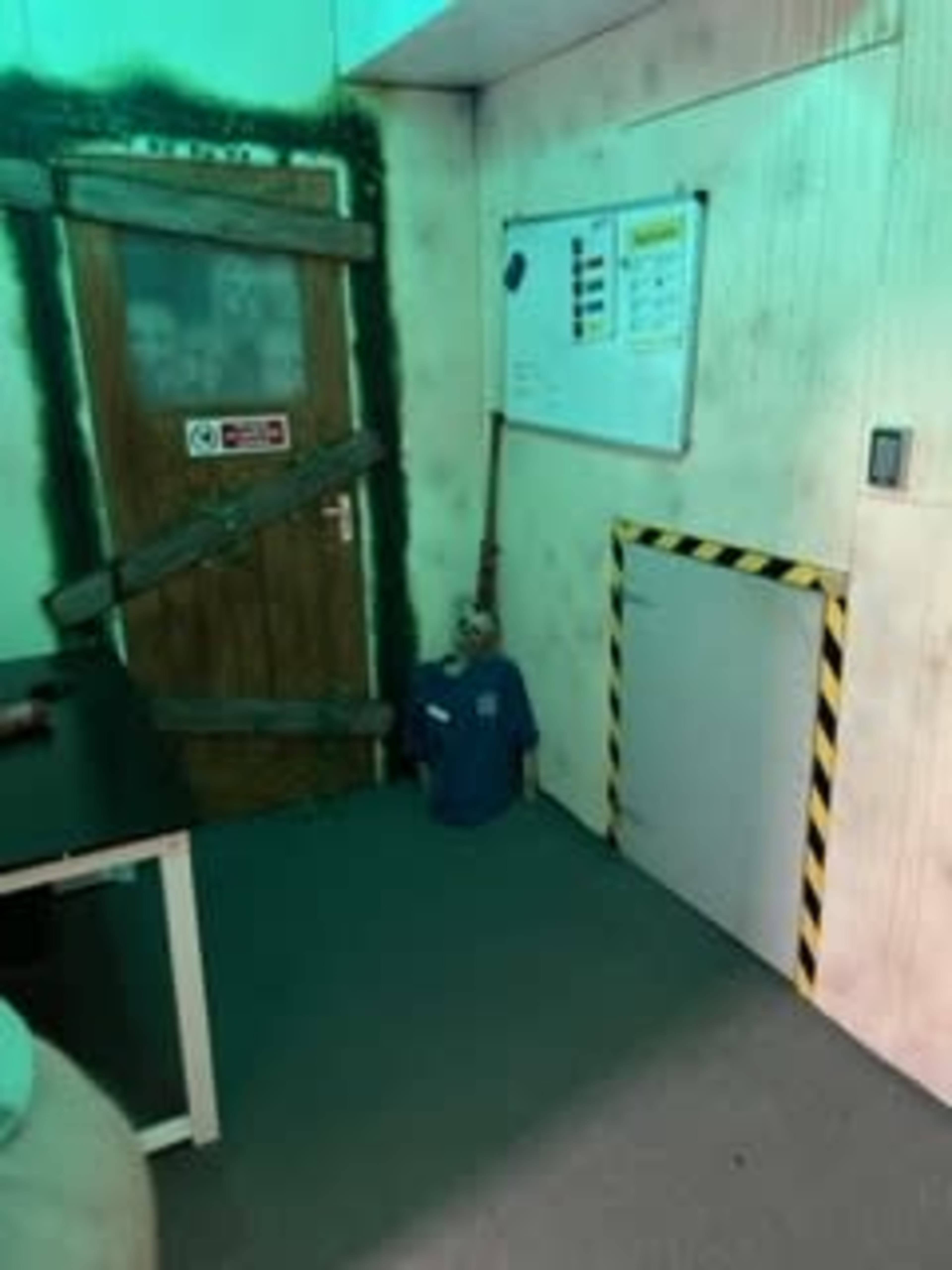 A room with a door that is boarded up, a small table, a bulletin board, and an unusual figure dressed in a blue shirt positioned in a corner.