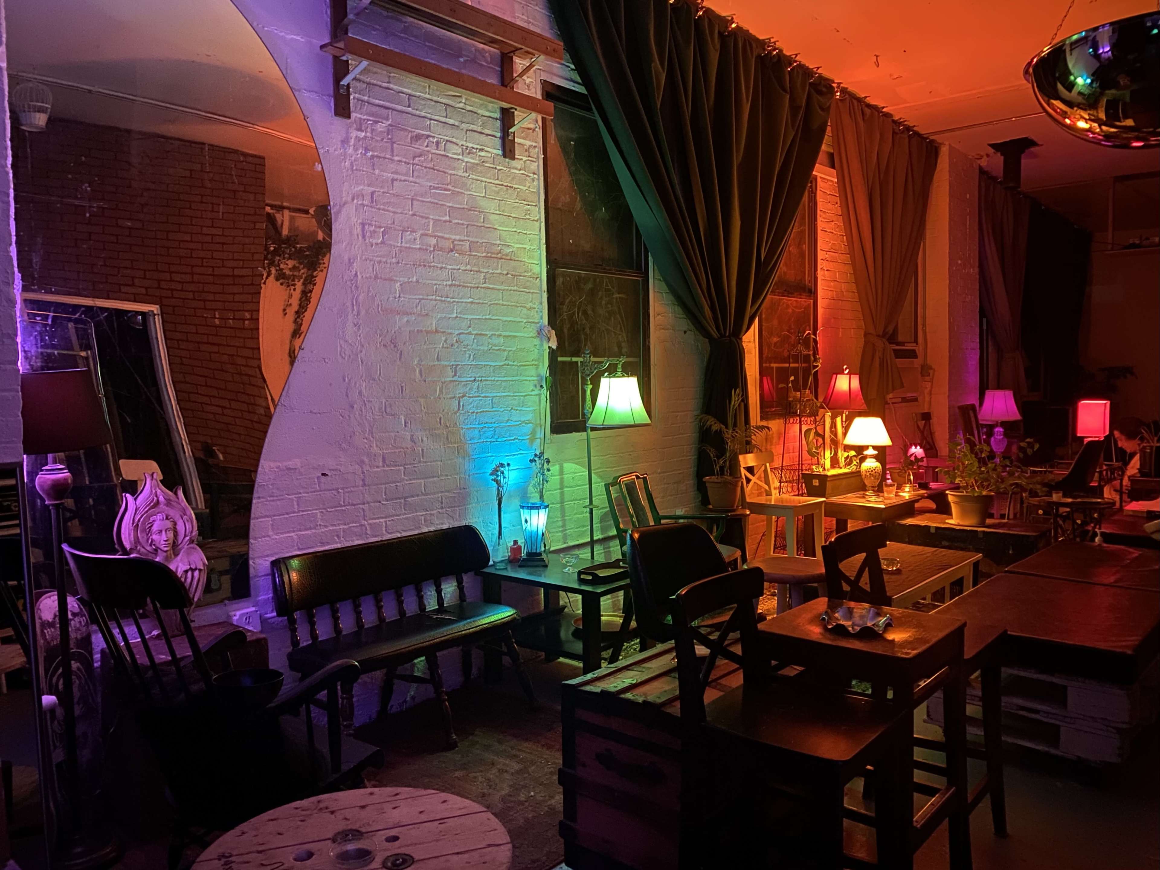 A dimly lit lounge area featuring colorful lighting, wooden furniture, and various lamps scattered throughout.