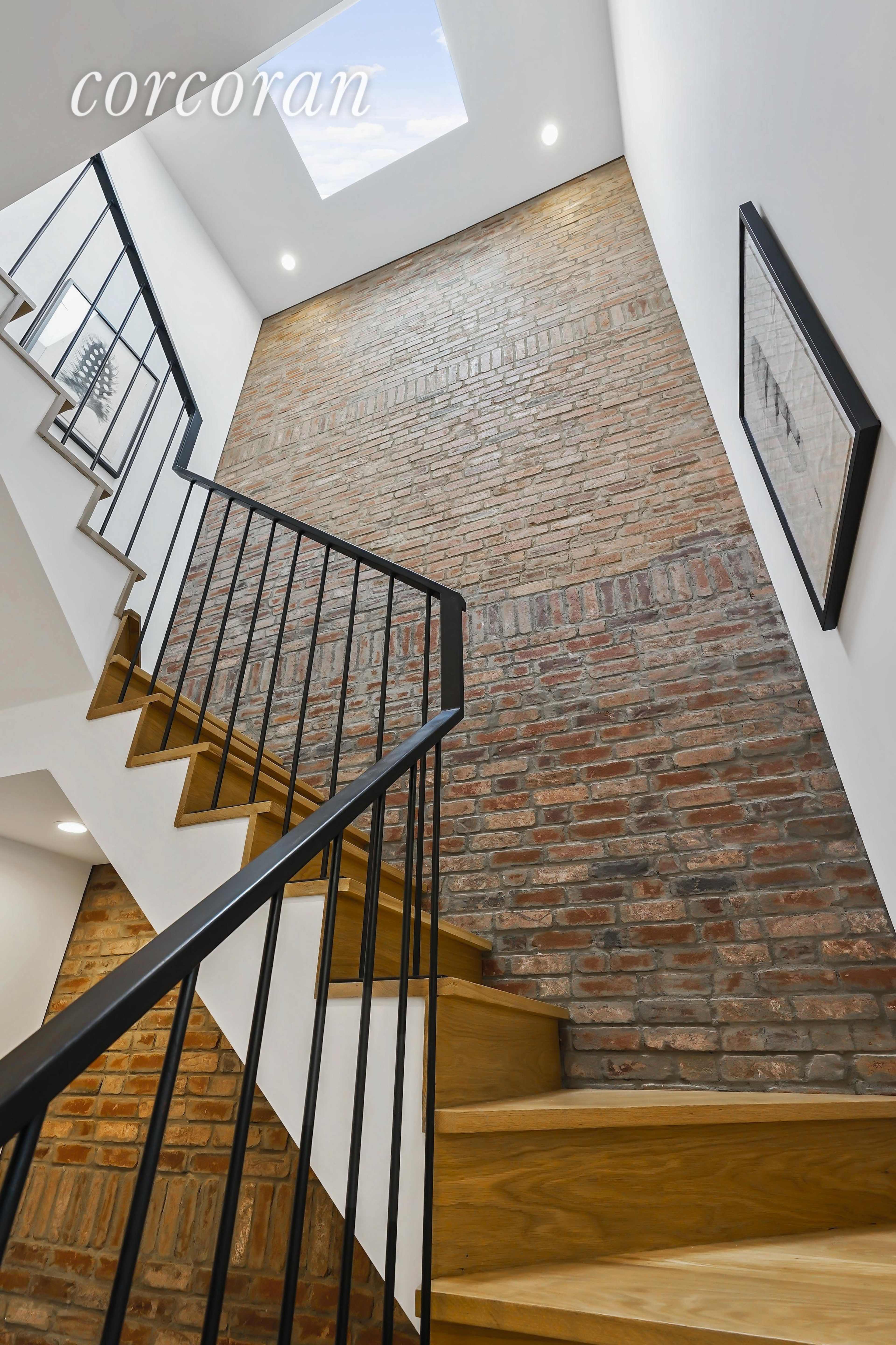 A staircase with wooden steps and a black railing ascends alongside a brick wall, leading to a skylight above.