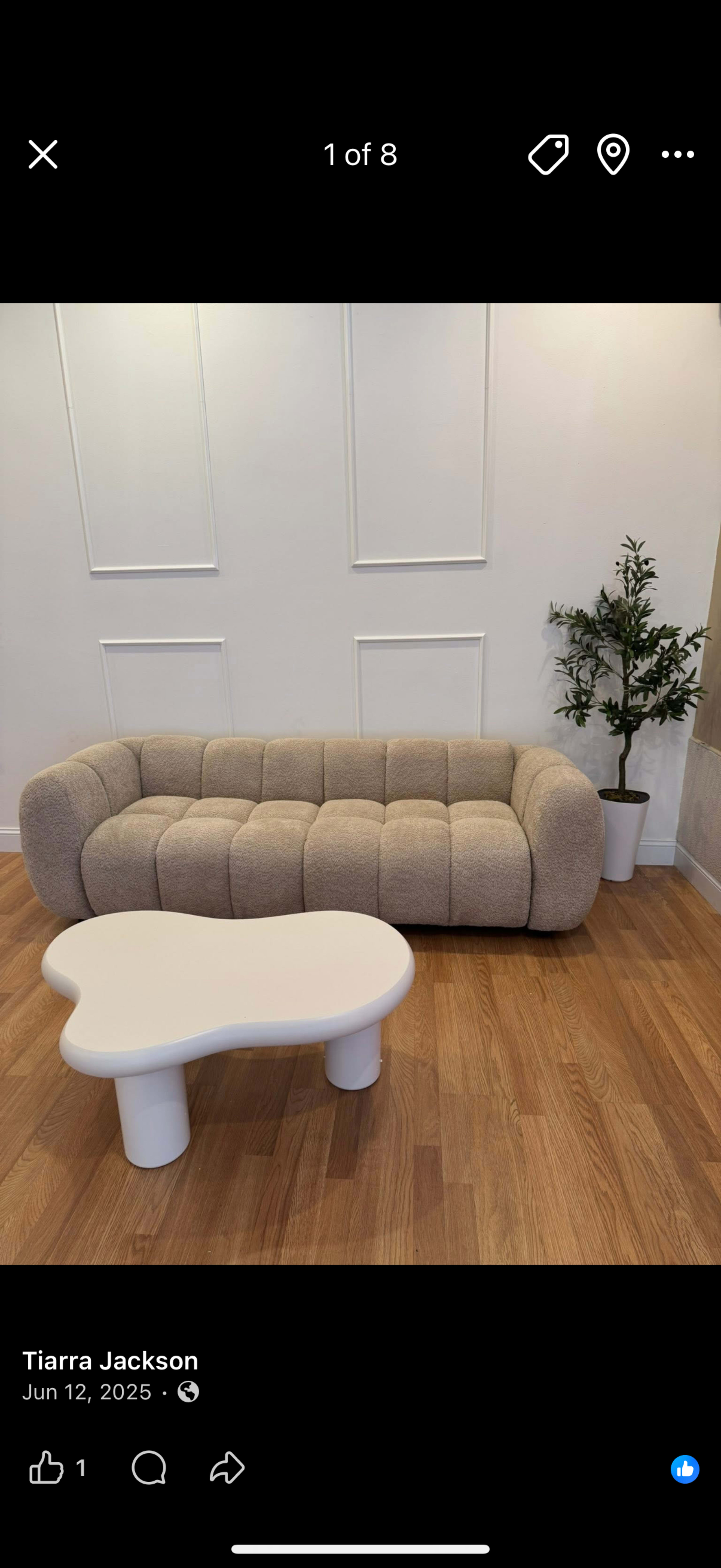 The image shows a curved beige couch and a unique white coffee table with rounded edges, positioned on a wooden floor against a plain white wall, along with a small green plant in the corner.