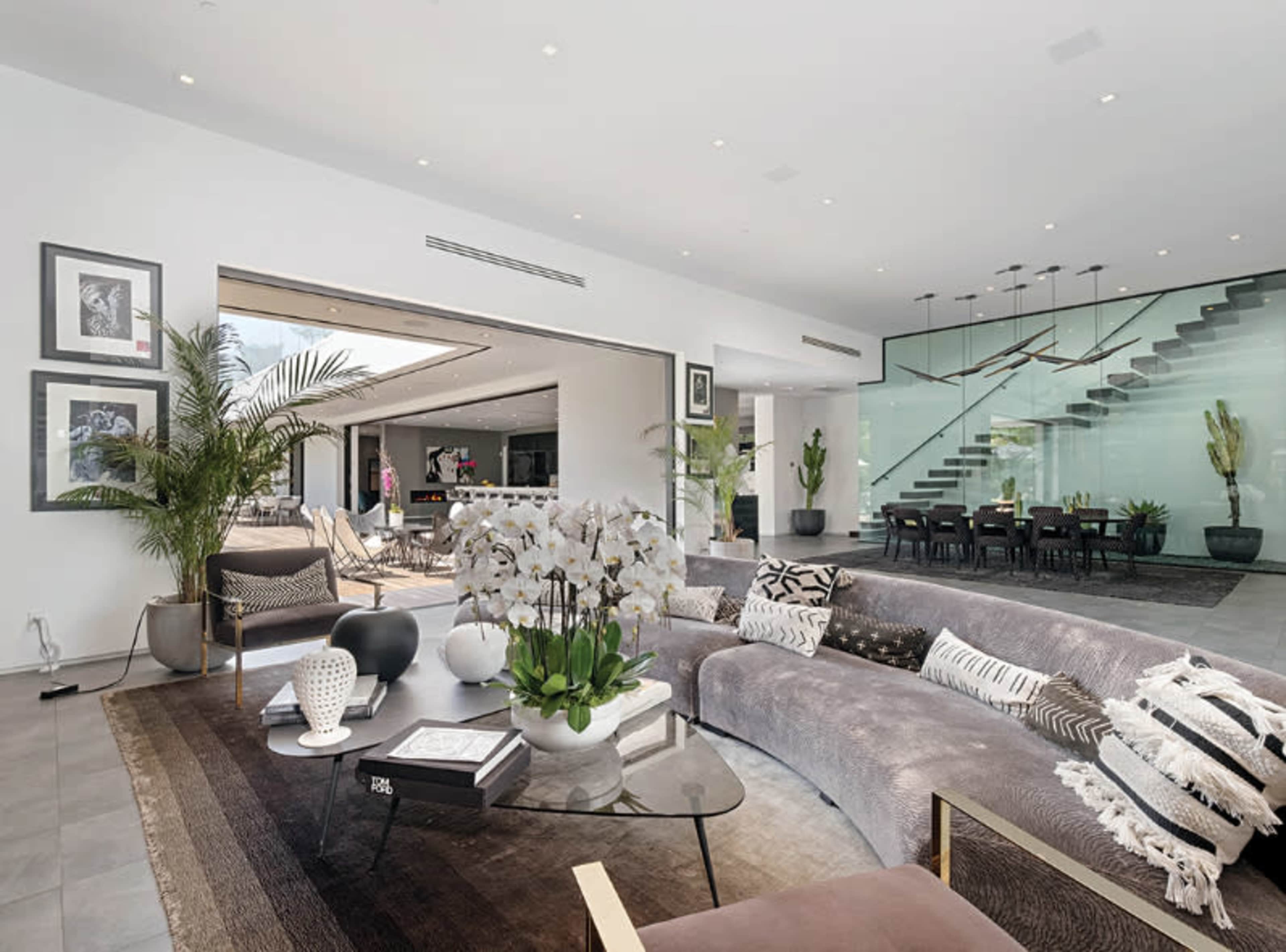 A modern living room features a large sectional sofa, a glass coffee table, and an open layout connecting to a dining area with stairs in the background.