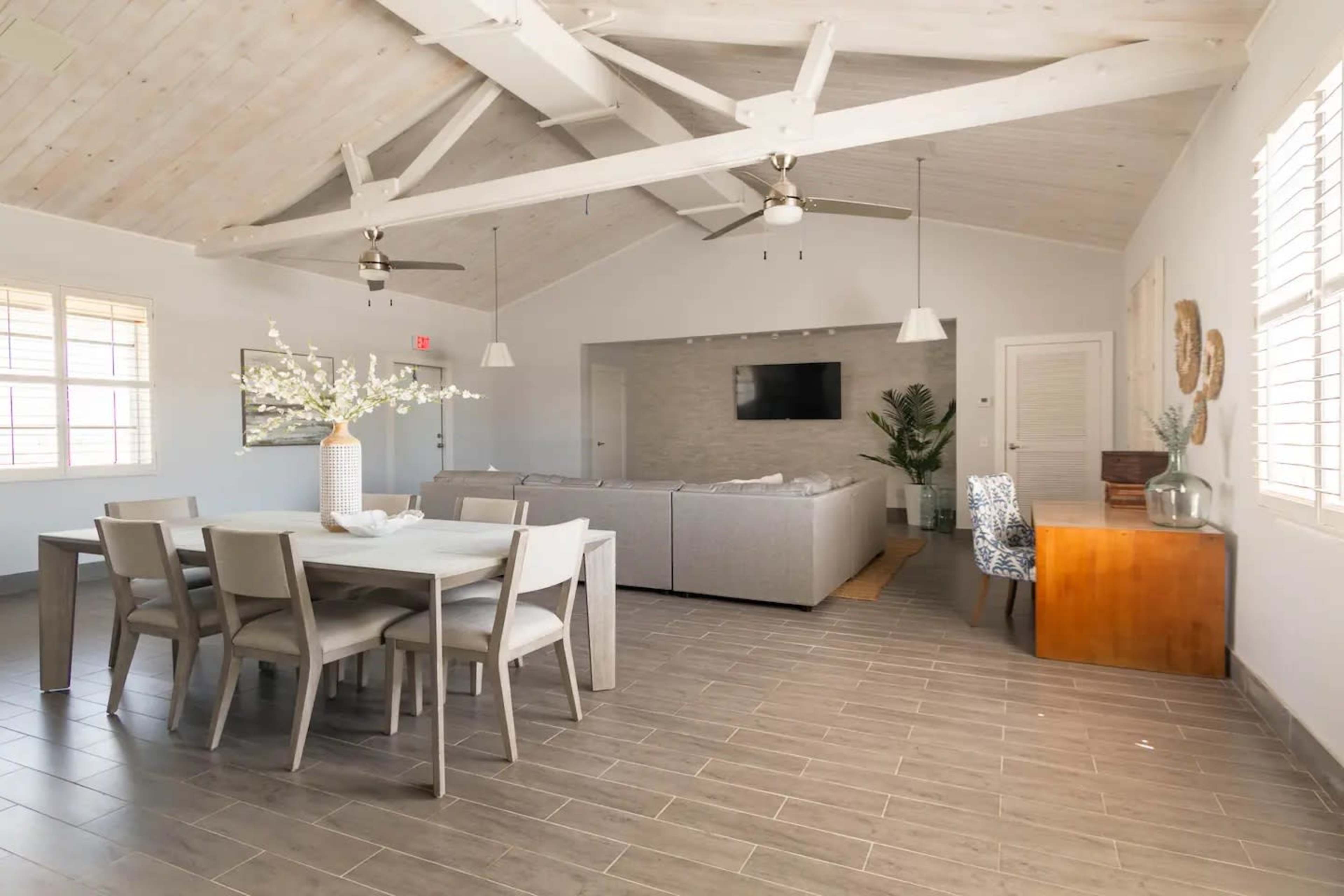 The image shows a bright, open-concept living space with a dining table, a large sectional sofa, and a wooden desk against a wall.