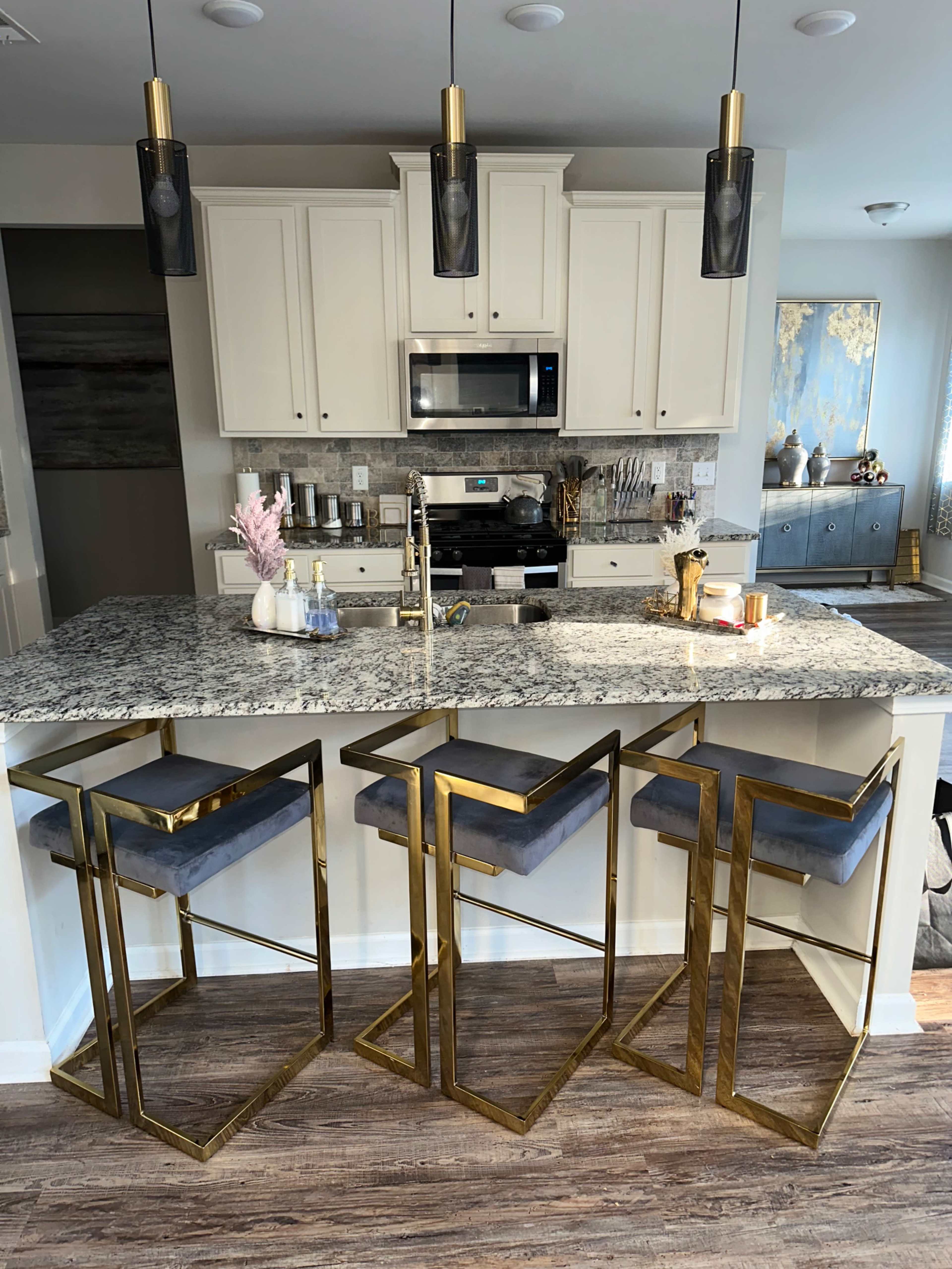 A modern kitchen features a granite countertop with three gold-framed bar stools and pendant lights overhead.