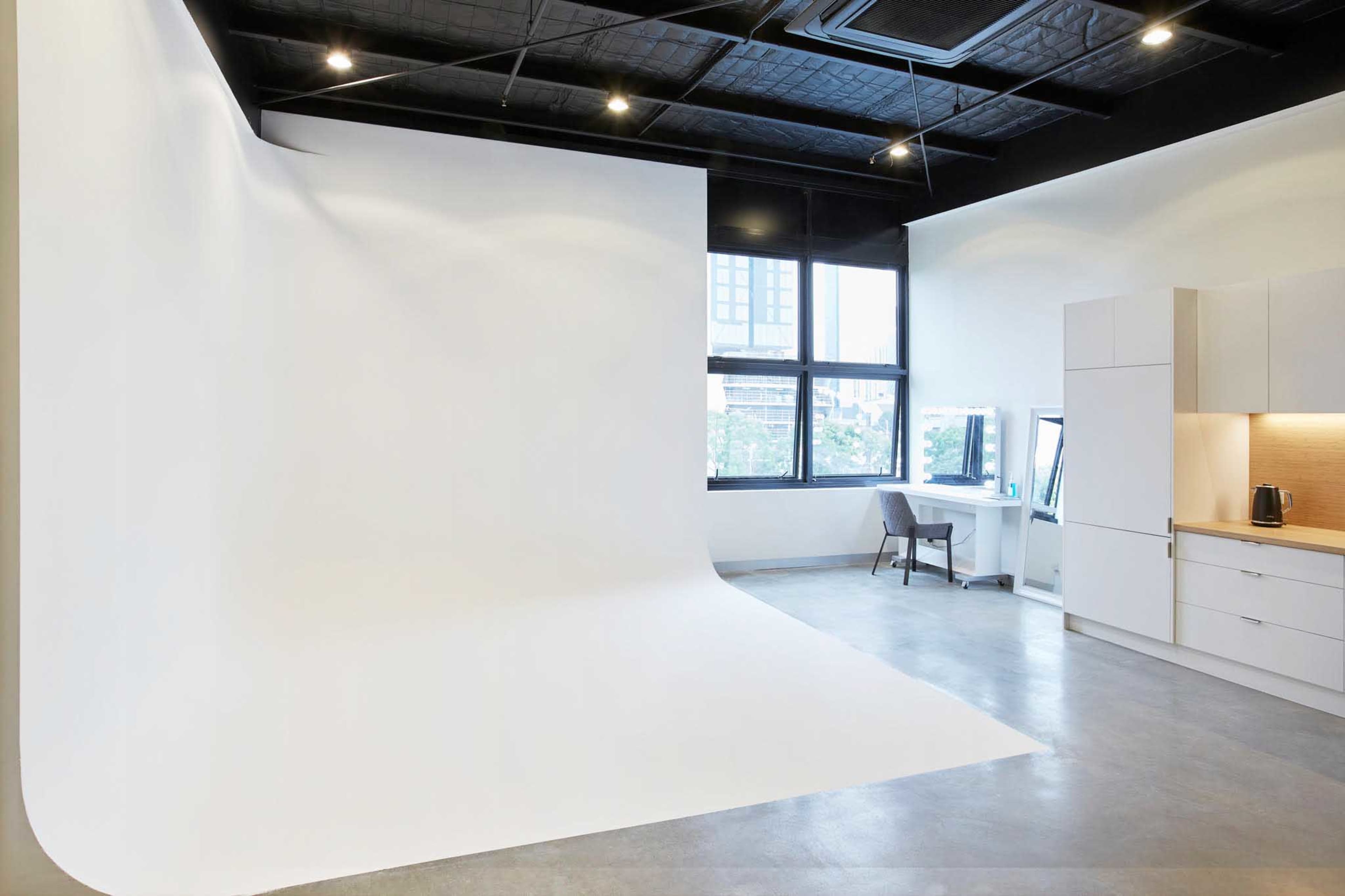 A minimalistic photography studio with a large white backdrop, a desk, and a kitchenette in a brightly lit room.
