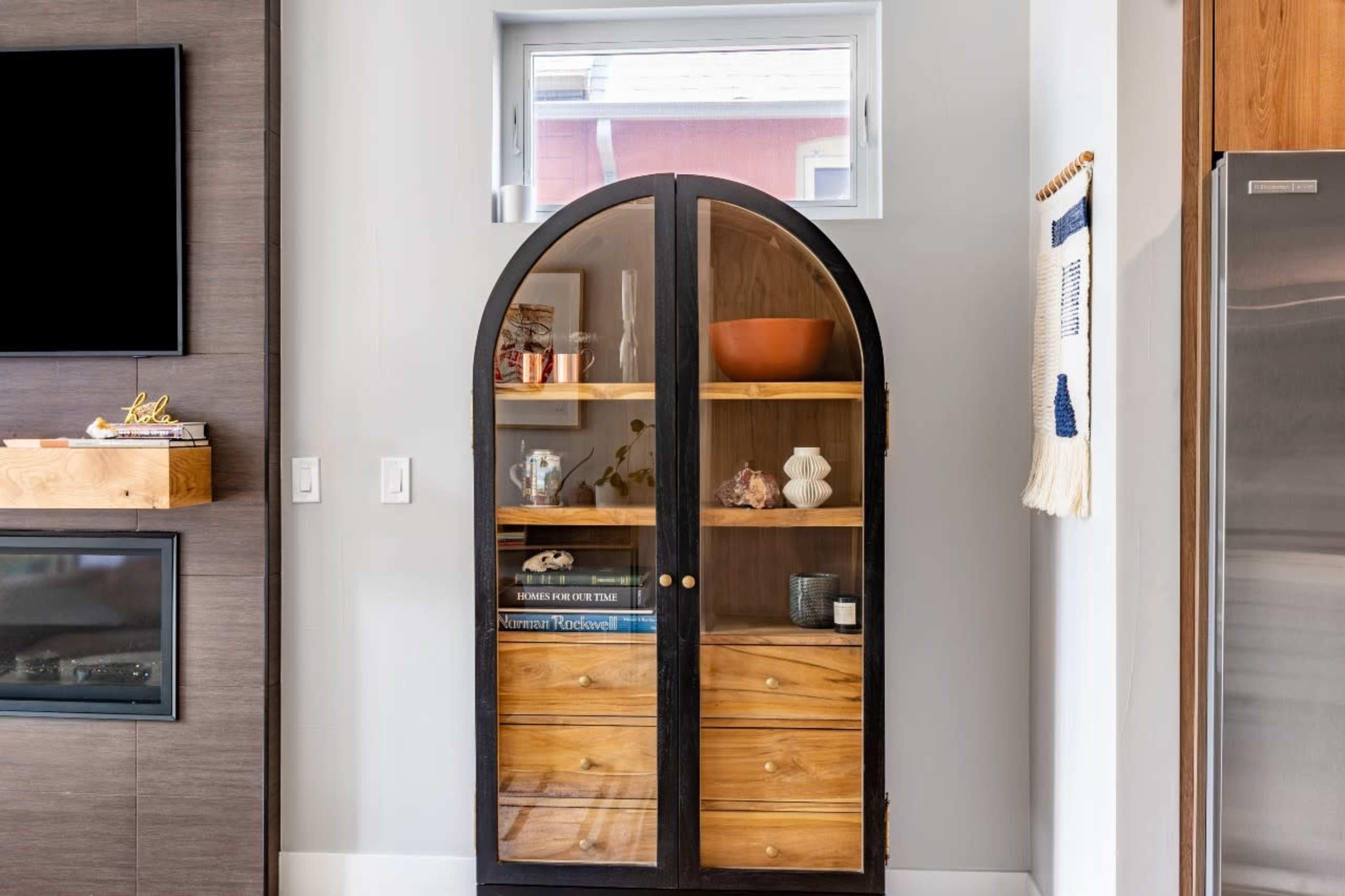 A wooden cabinet with glass doors displays various items, positioned against a wall beside a television and a silver refrigerator.