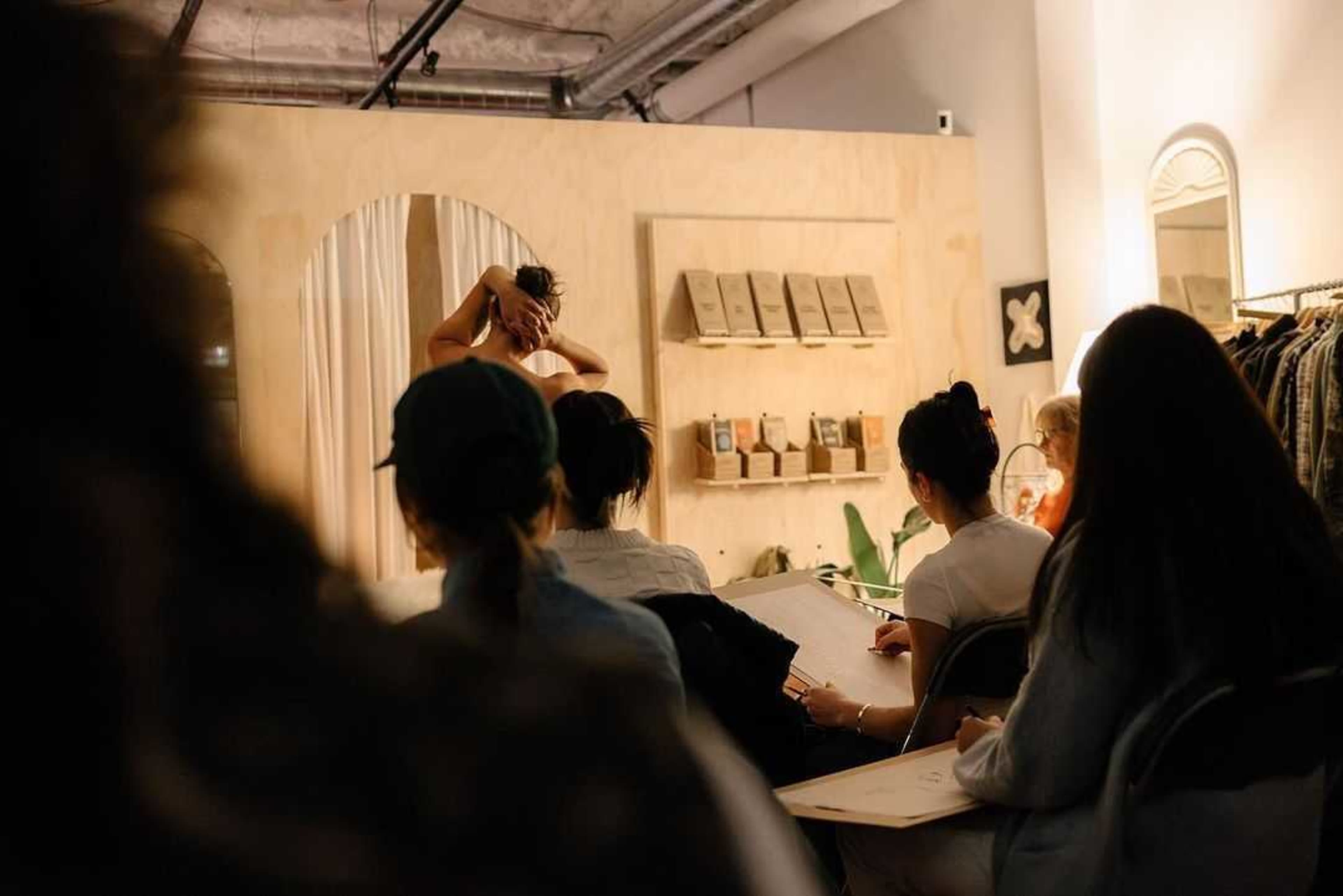 A group of people sketching a model posing in a studio setting, with several design elements and displays in the background.