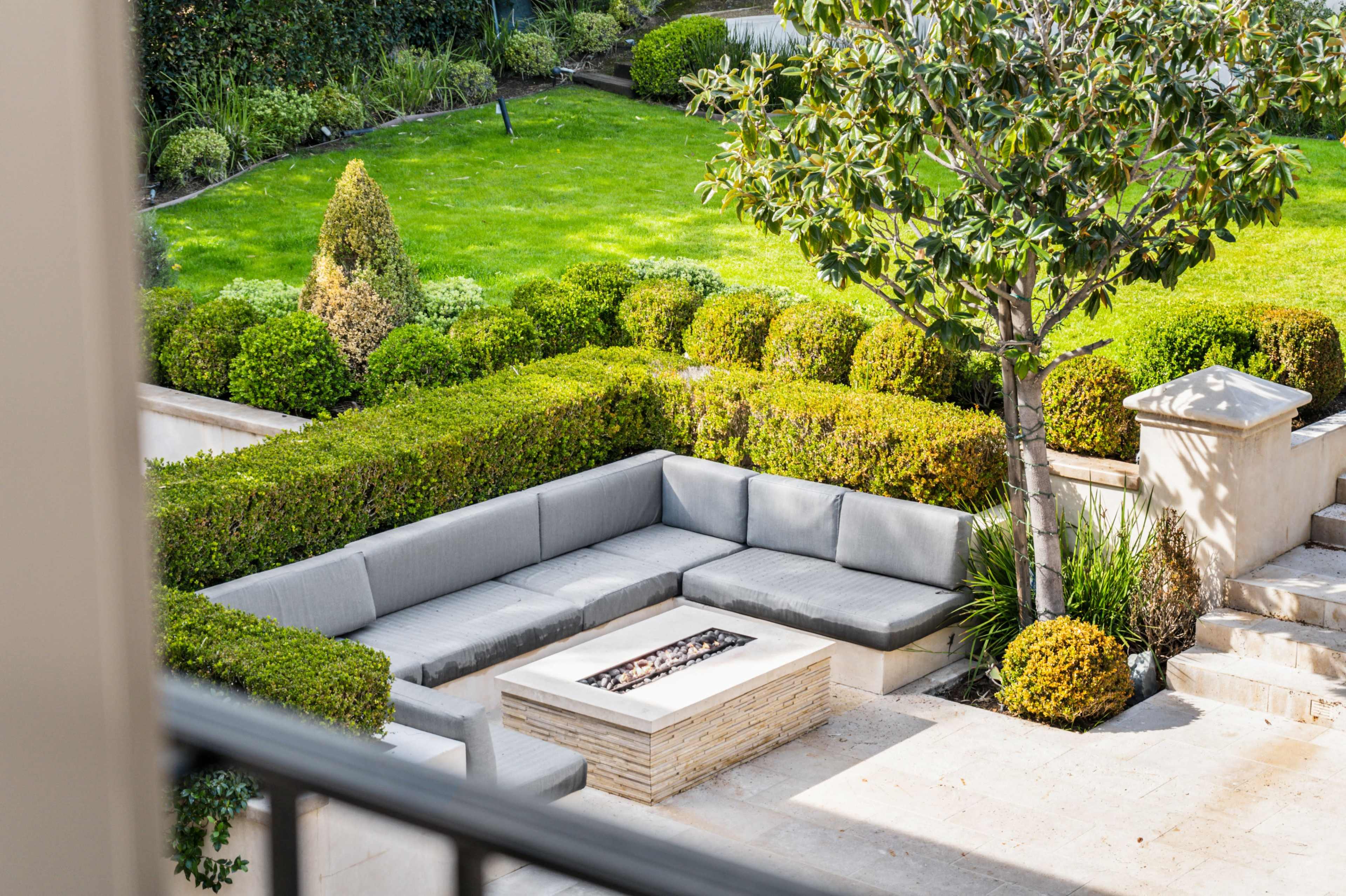 A modern outdoor seating area with gray sectional sofas surrounding a stone fire pit, set against neatly trimmed hedges and a green lawn.