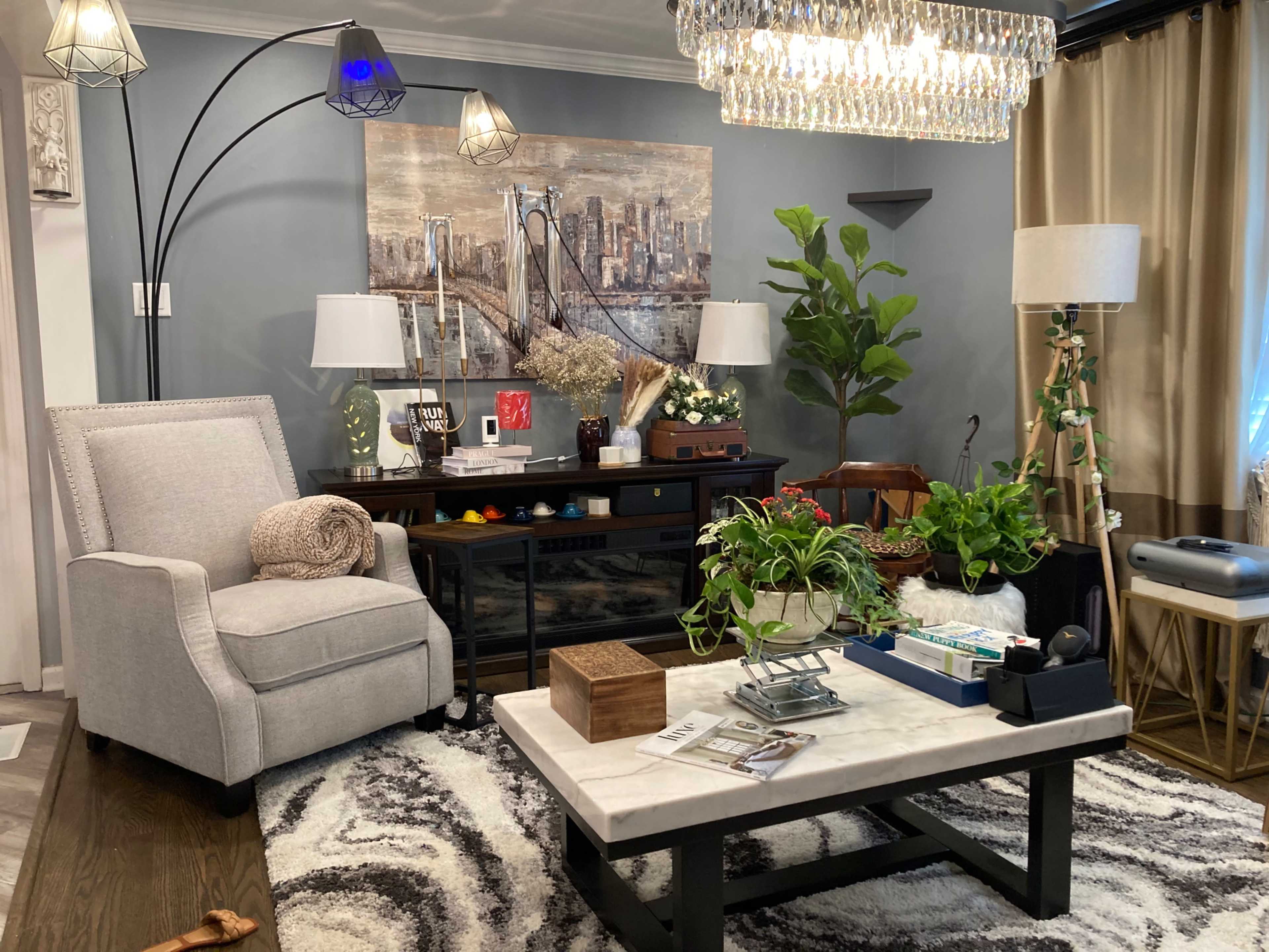 A modern living room with a gray armchair, a coffee table with a marble top, various indoor plants, and decorative wall art featuring a city skyline.