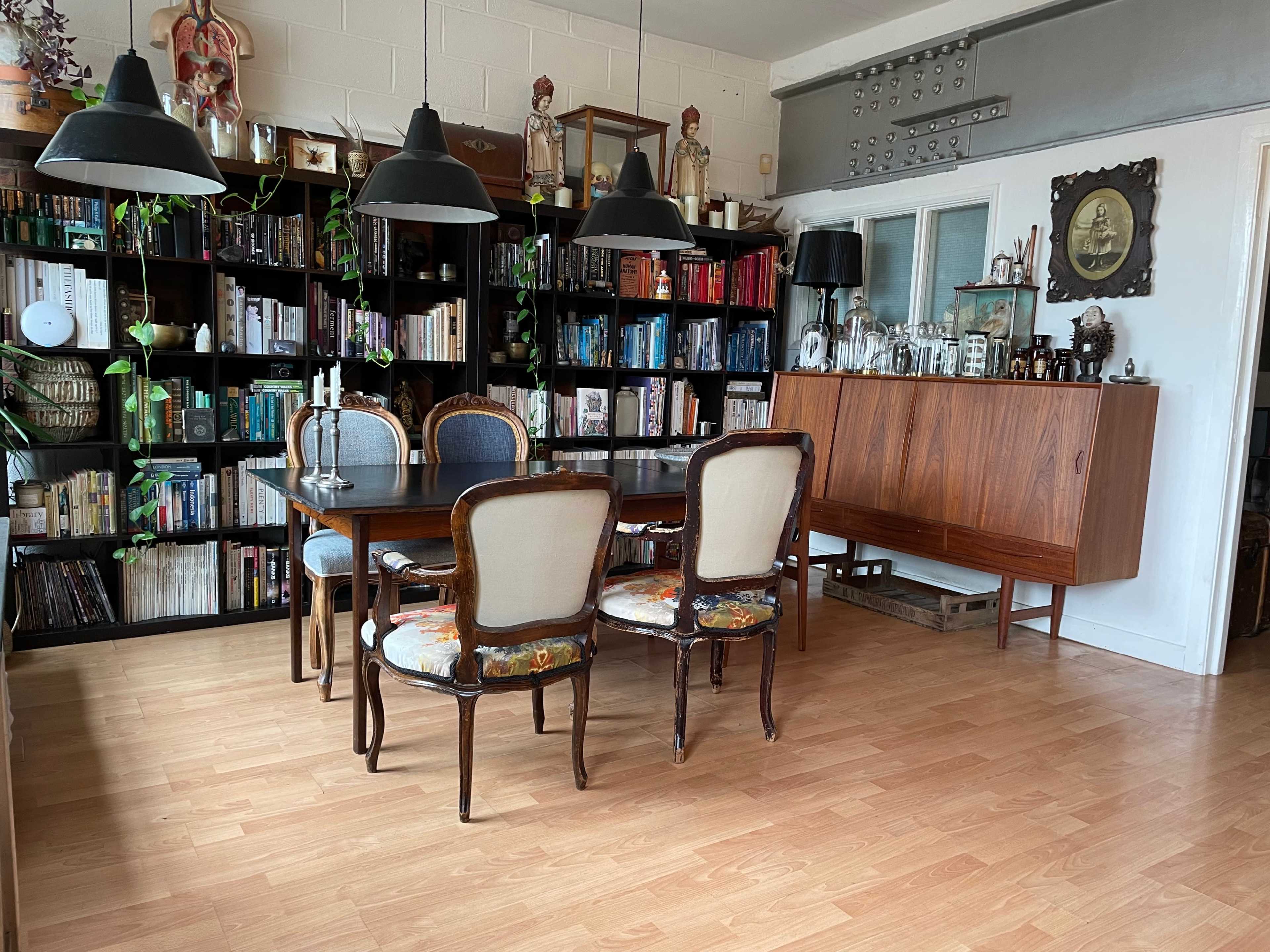 A dining area with a wooden table surrounded by four chairs, a bookshelf filled with books and plants, and a sideboard against the wall.
