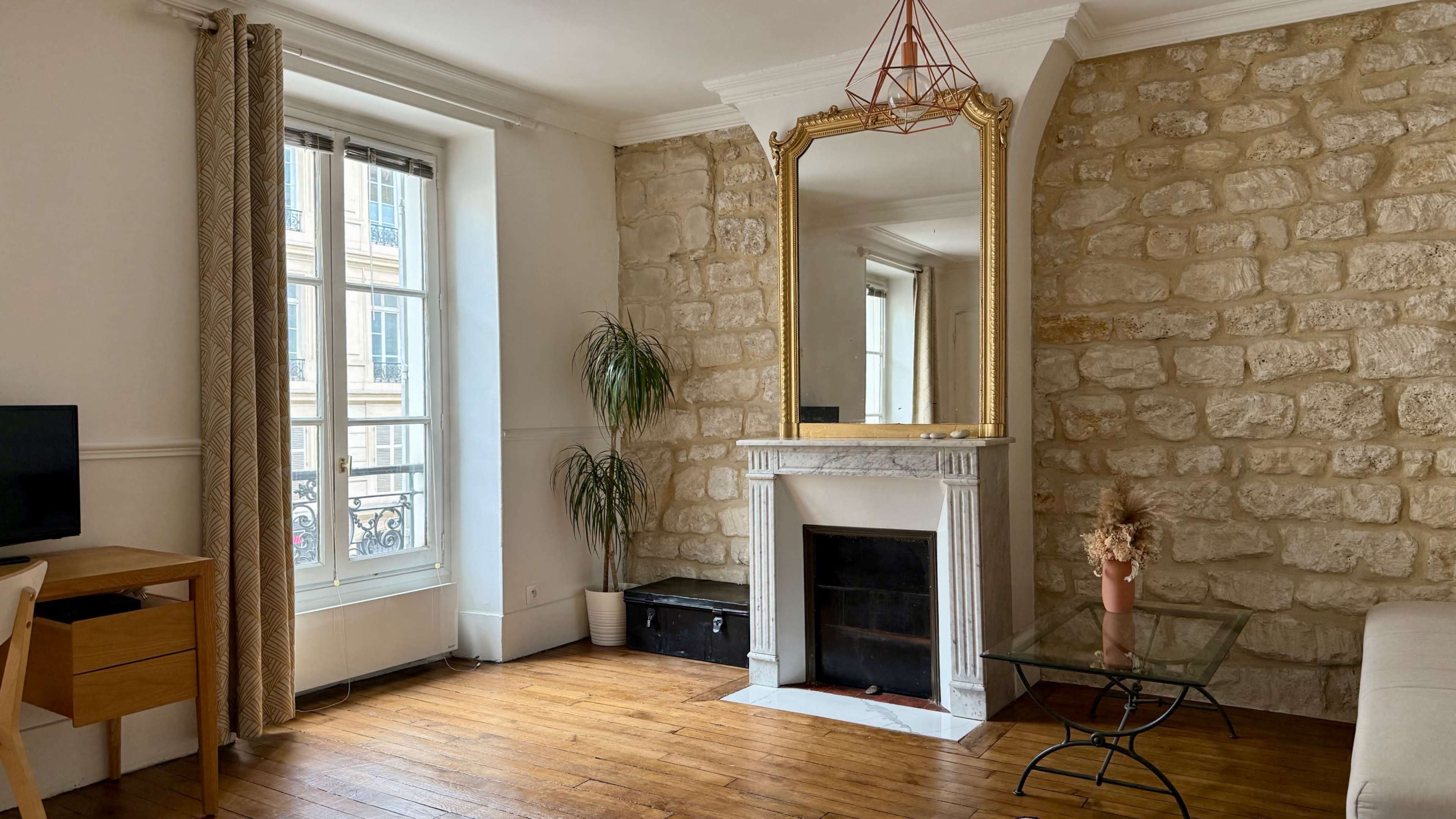 The image shows a cozy living room with a stone wall, a large mirror above a marble fireplace, a window with natural light, and wooden flooring.