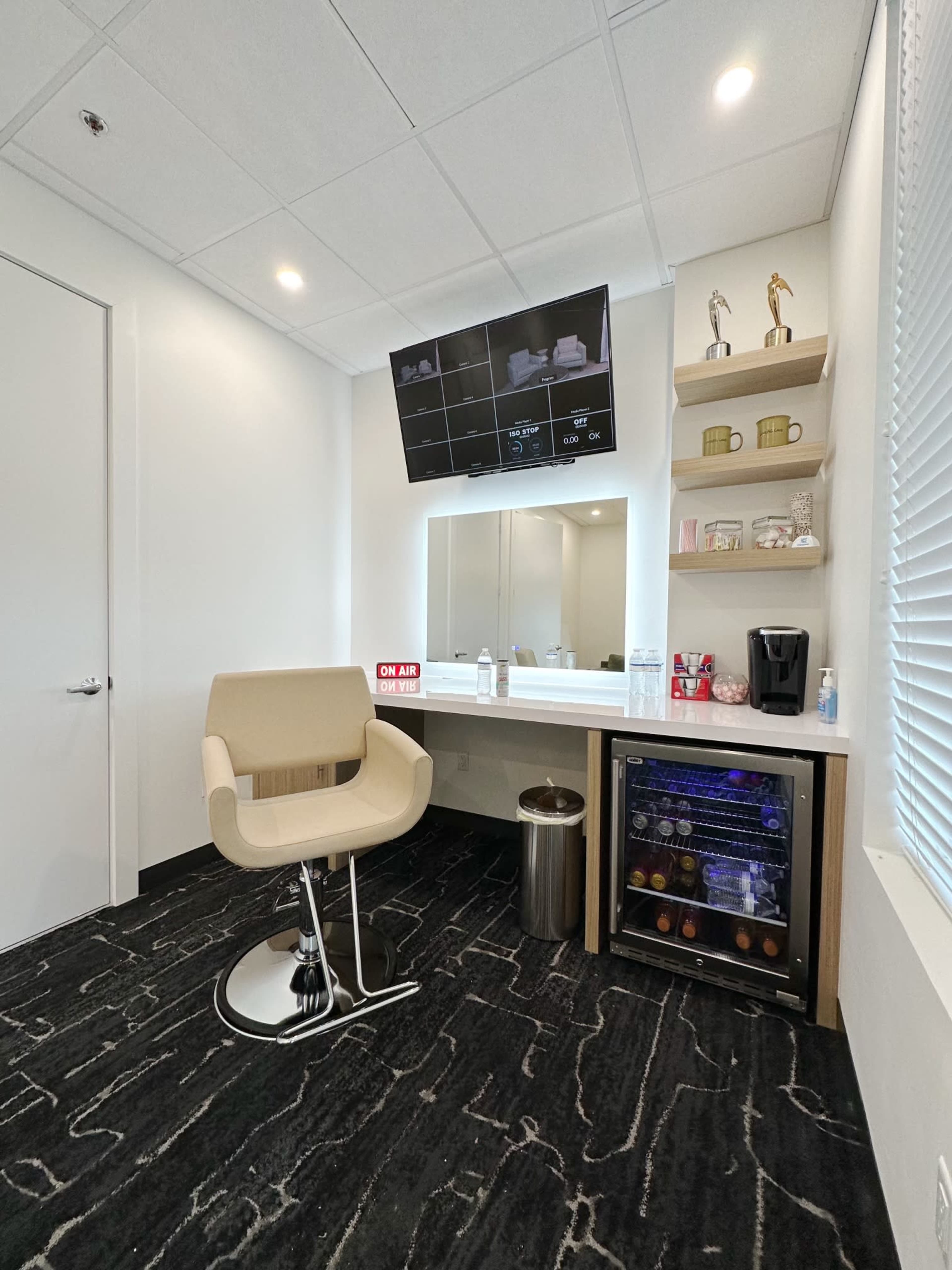 A small room featuring a vanity mirror, a modern chair, a refrigerator, and a TV mounted on the wall.