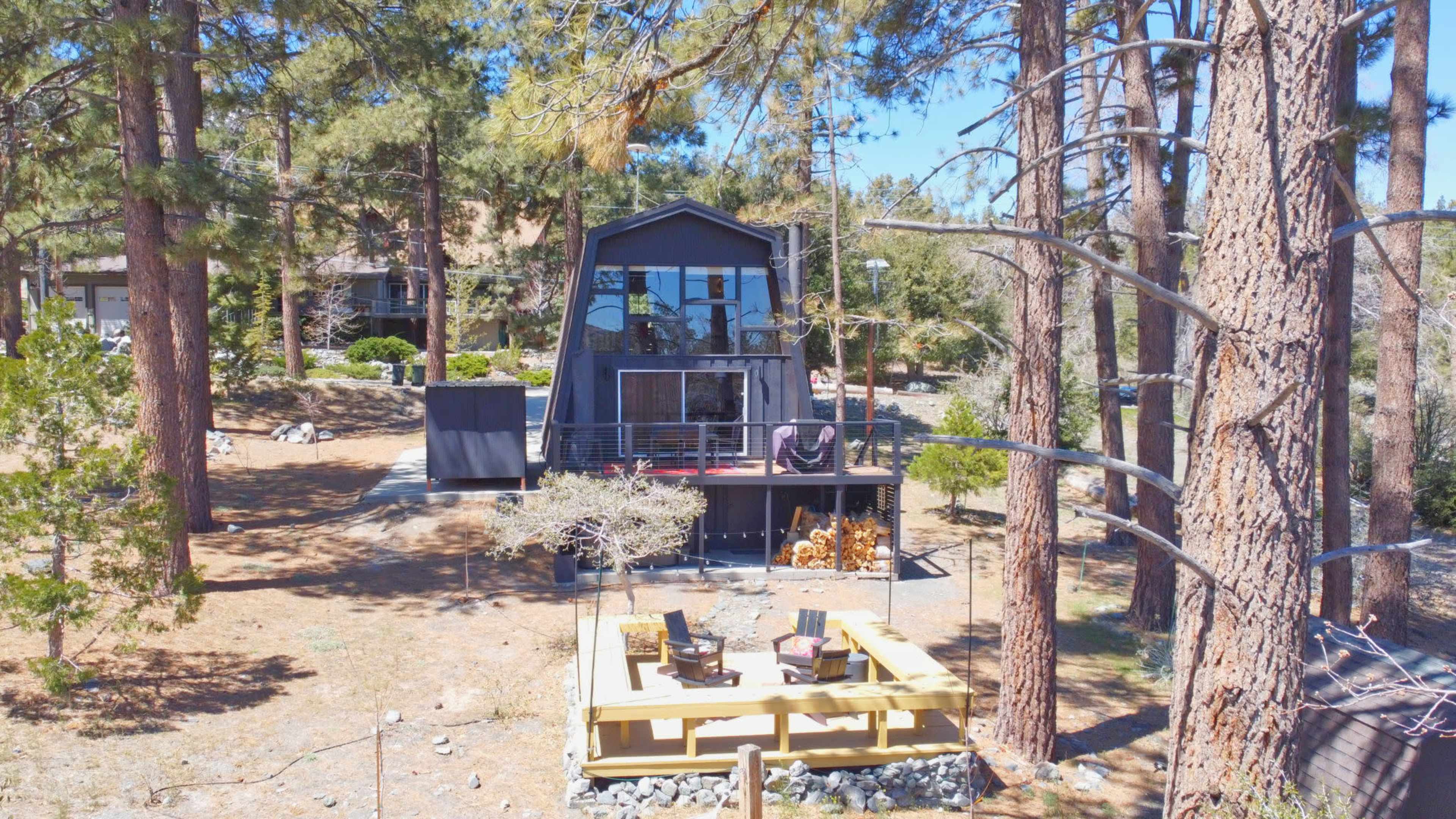 A modern two-story black house with a deck is surrounded by tall pine trees and features a patio area with seating in the foreground.