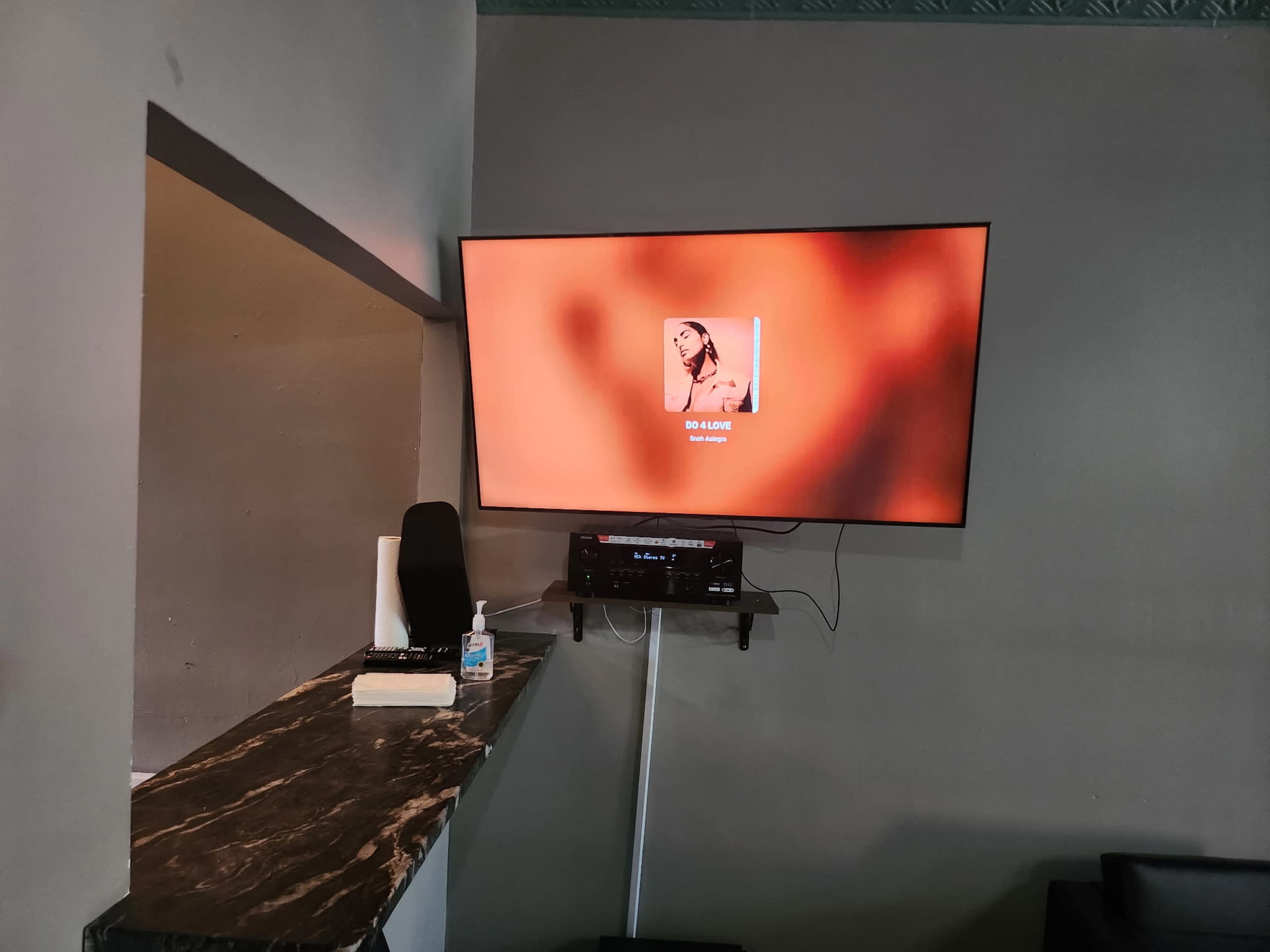 A large television displaying a music artist's photo is mounted on a gray wall near a marble-topped bar area.