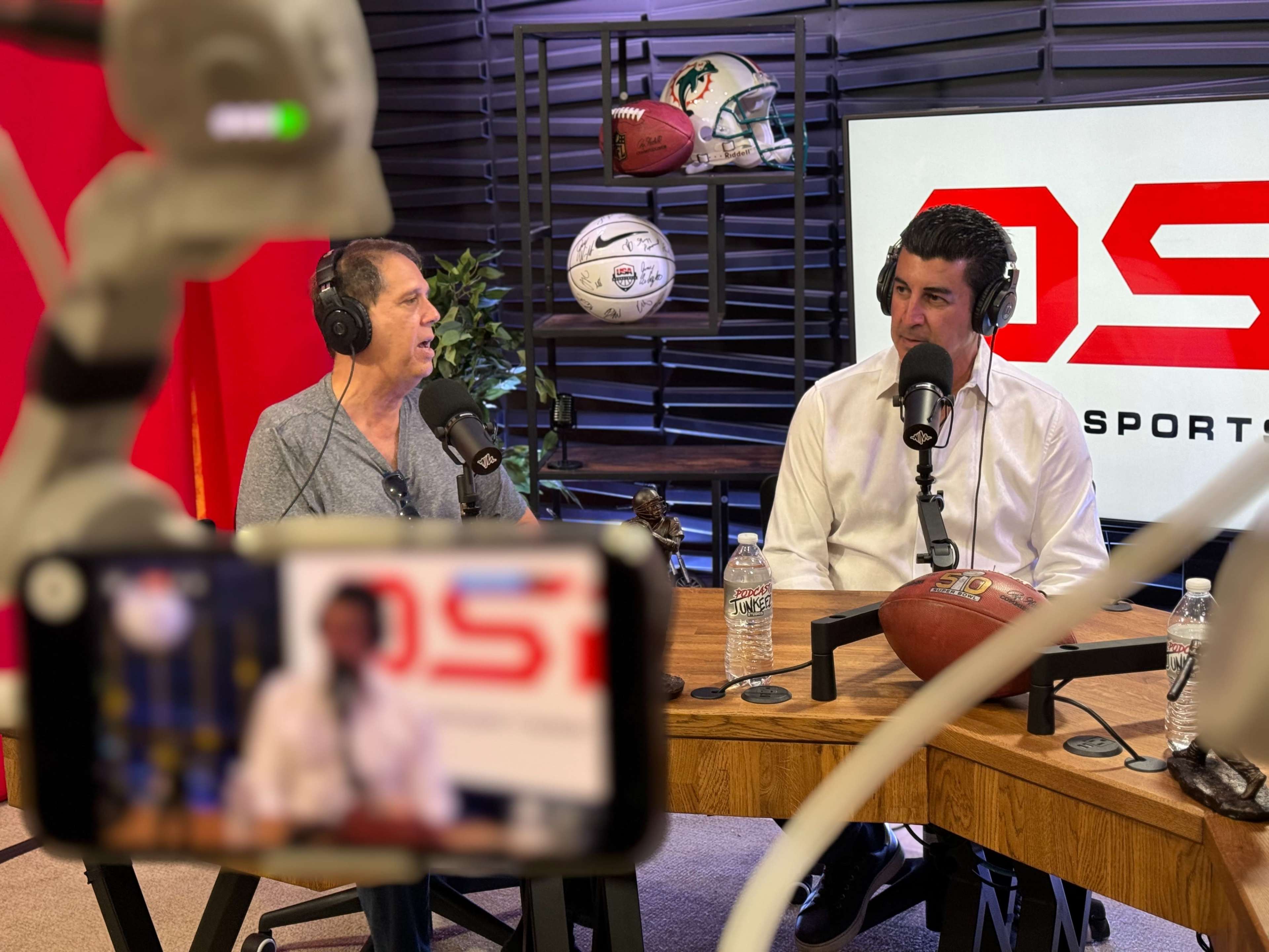 Two men are sitting at a wooden table, speaking into microphones in a studio with sports memorabilia in the background.