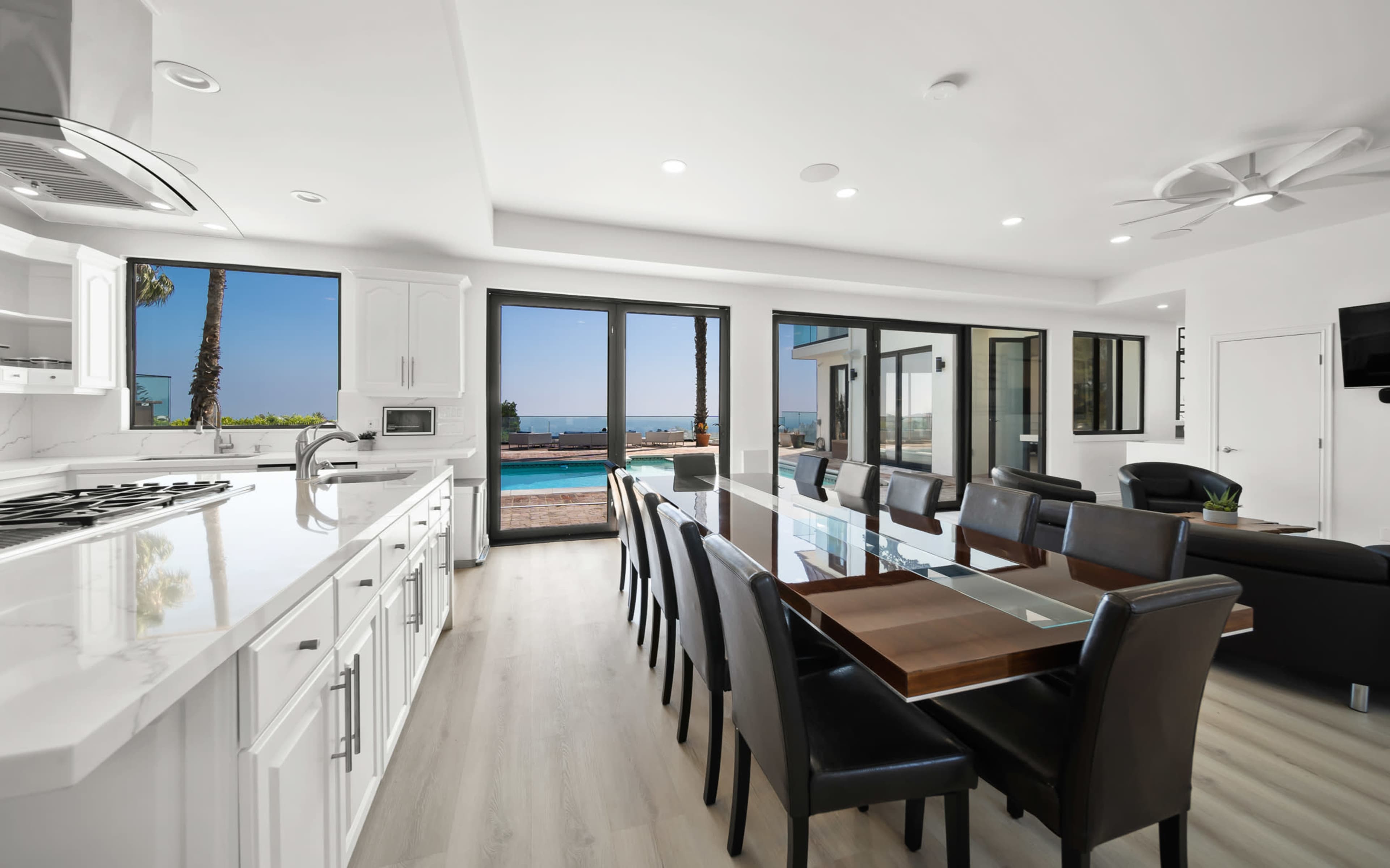 A modern kitchen and dining area features a large table surrounded by black chairs, with sliding glass doors that open to a patio and ocean view.