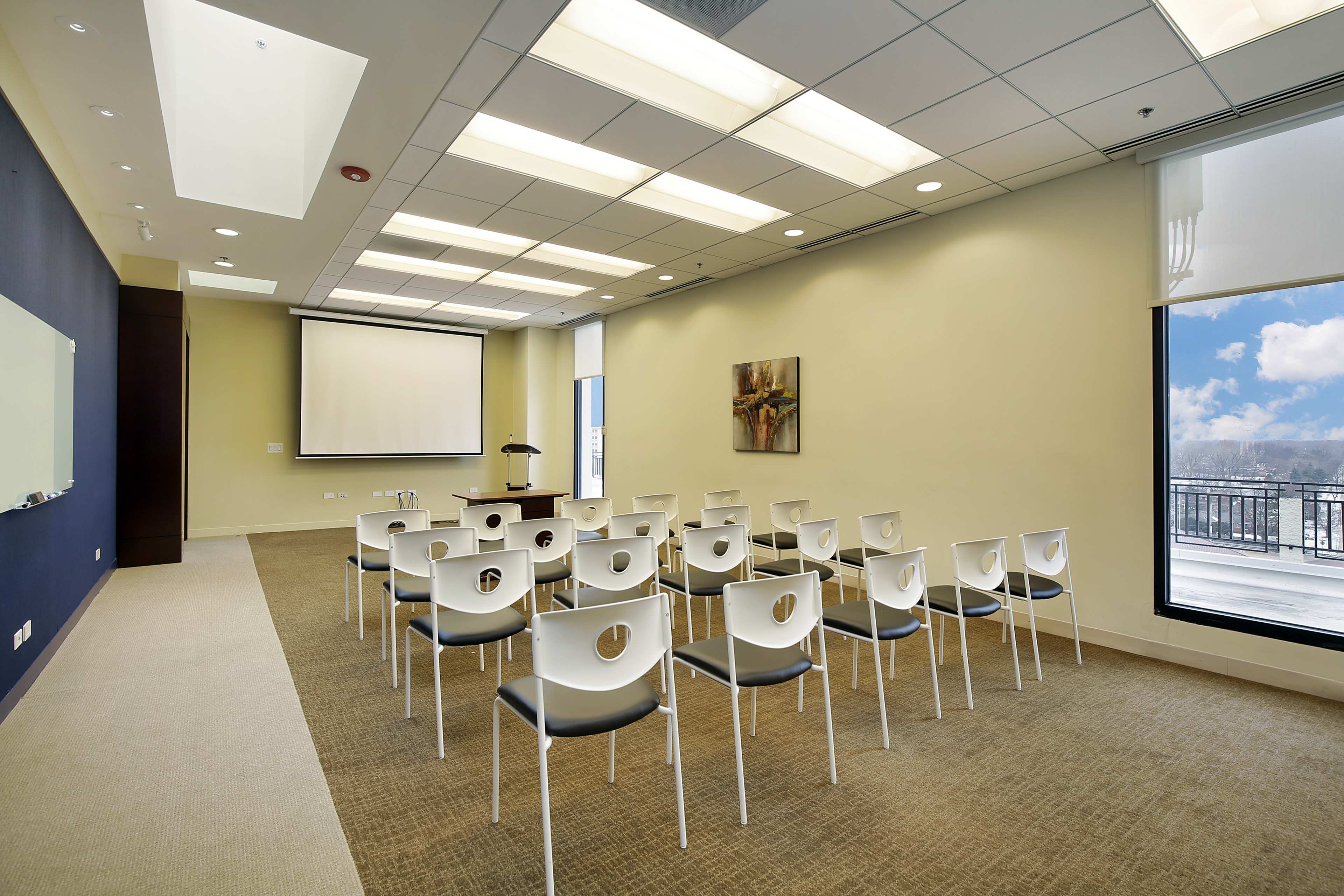 A meeting room is set up with rows of white chairs facing a projector screen and a podium, with large windows letting in natural light.