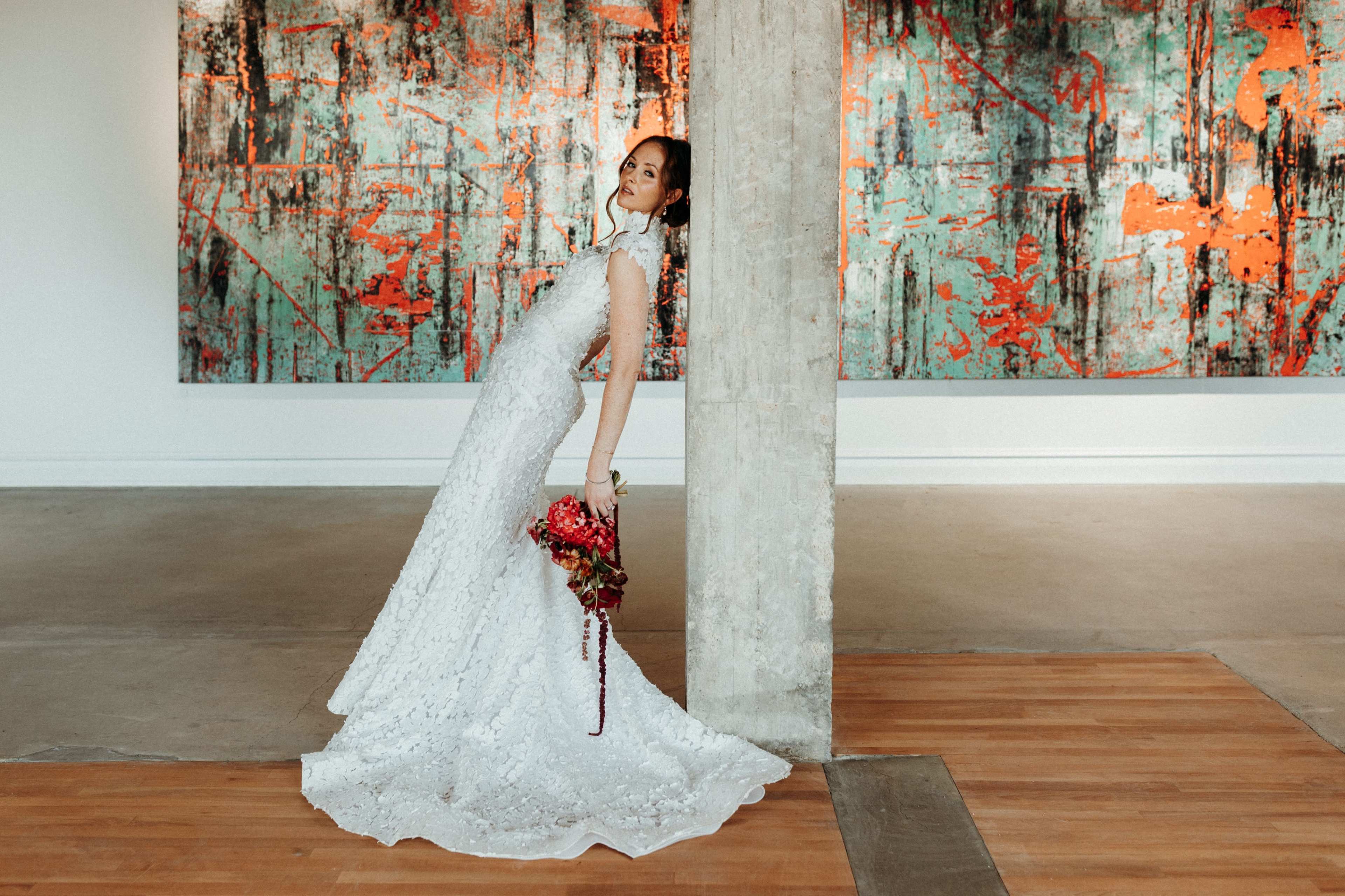 A bride in a white lace dress poses elegantly beside a concrete pillar in an art gallery featuring vibrant abstract paintings.