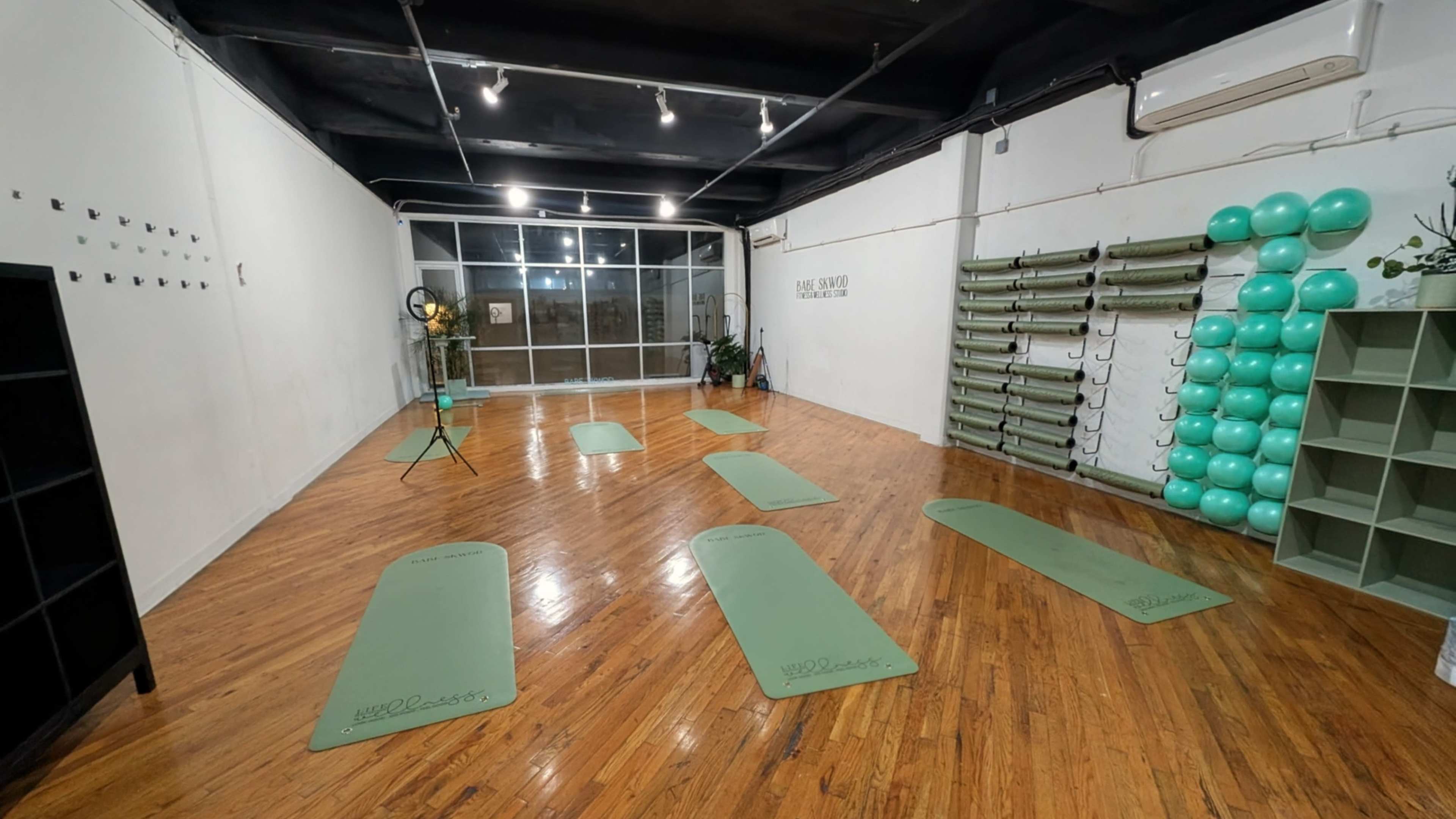 The image shows a yoga studio with wooden flooring and several green yoga mats arranged in a semi-circle, along with fitness equipment stored on the wall.