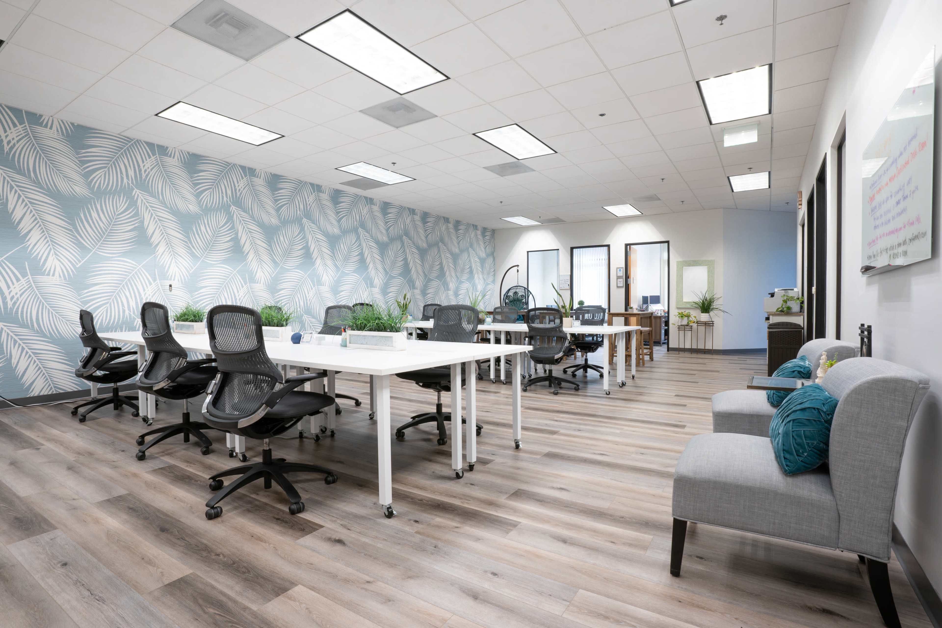 The image shows a modern office space with several workstations, stylish chairs, and a blue leafy-patterned accent wall.
