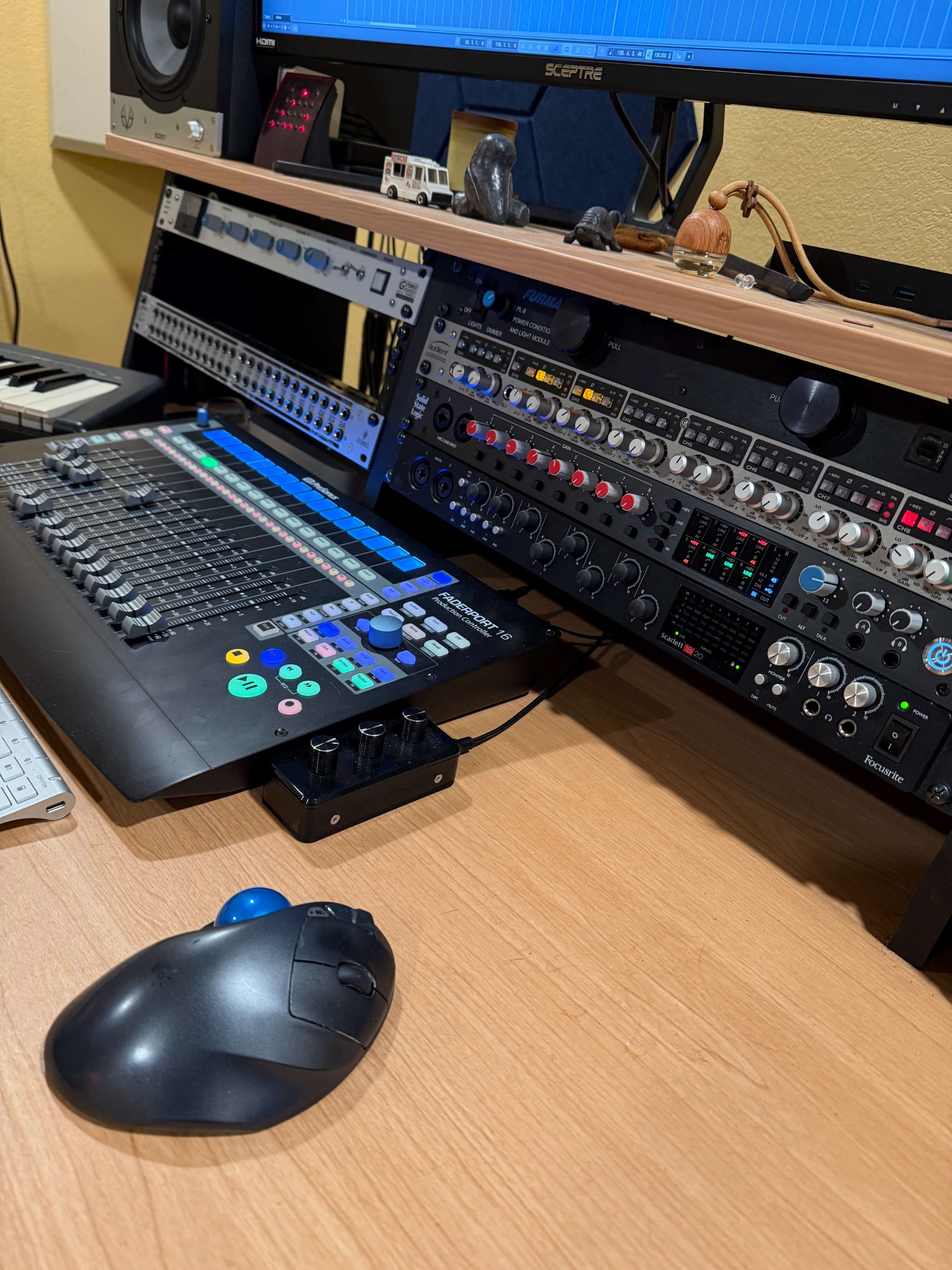 The image shows a music production setup with a digital audio workstation, control surface, audio interfaces, and computer peripherals arranged on a wooden desk.