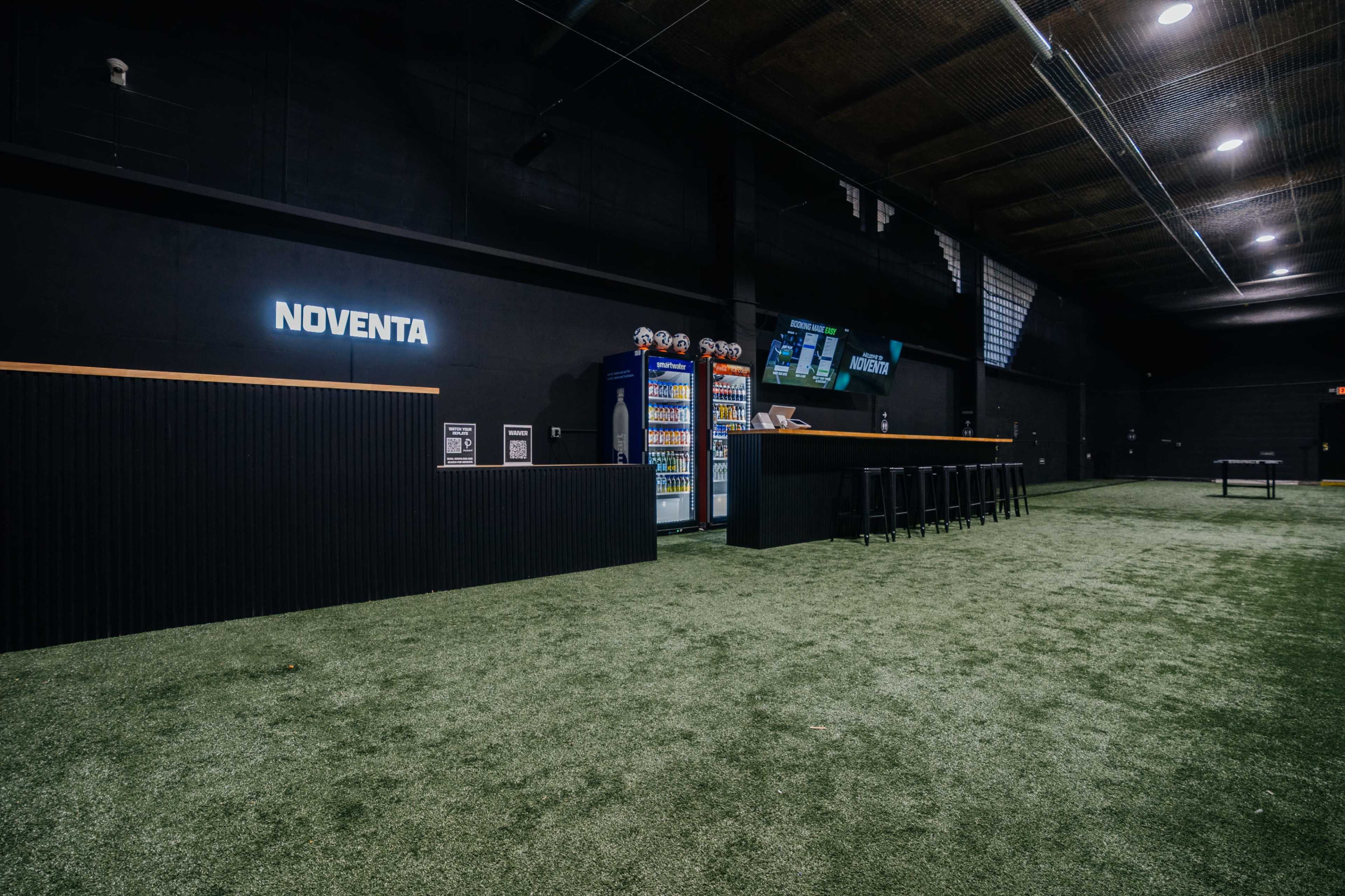 The image shows a modern indoor space with a dark color scheme, featuring a bar area with high stools, a beverage cooler, and a green artificial turf floor.