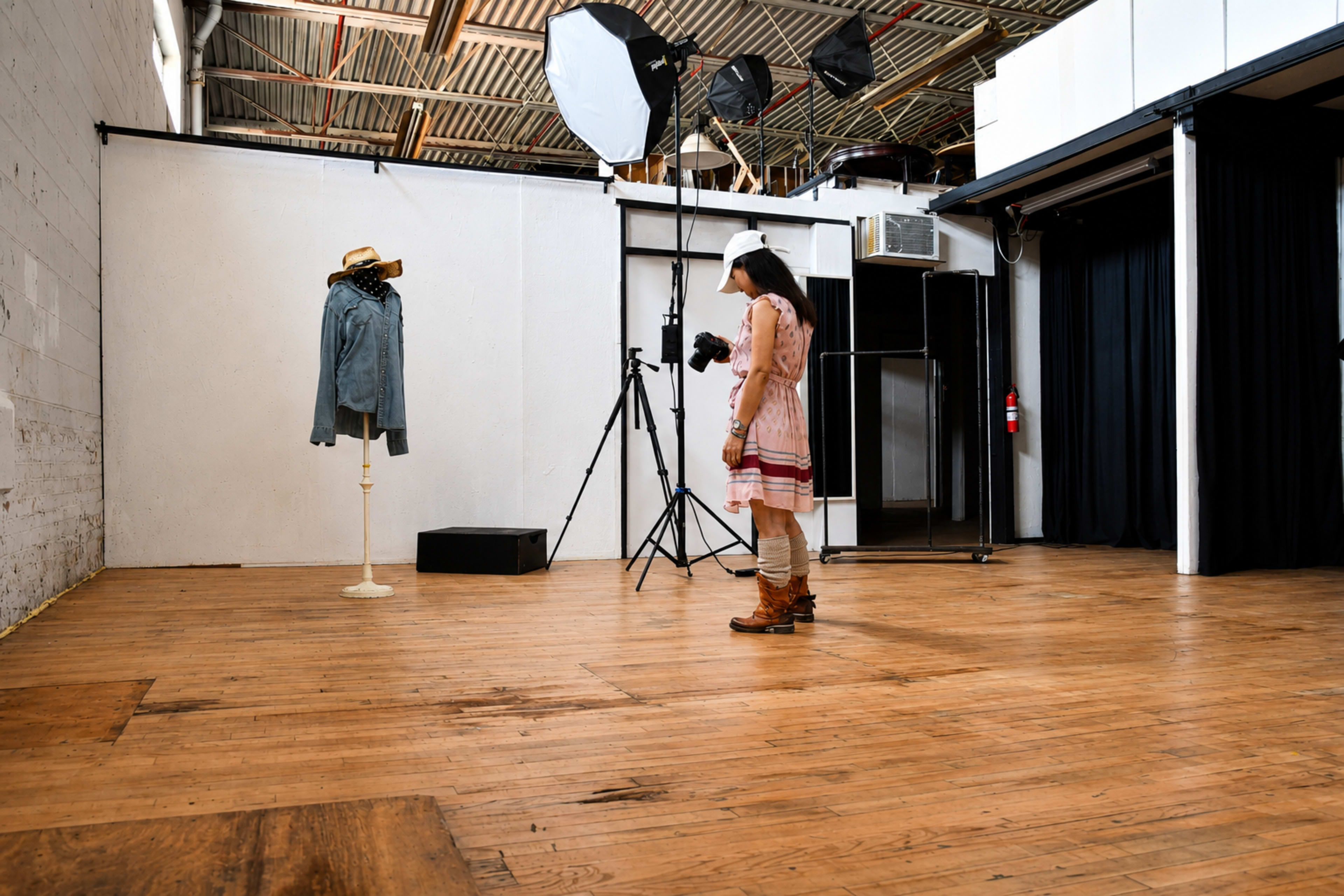 A young girl in a pink dress stands in a photography studio, facing a mannequin dressed in a denim jacket and hat.