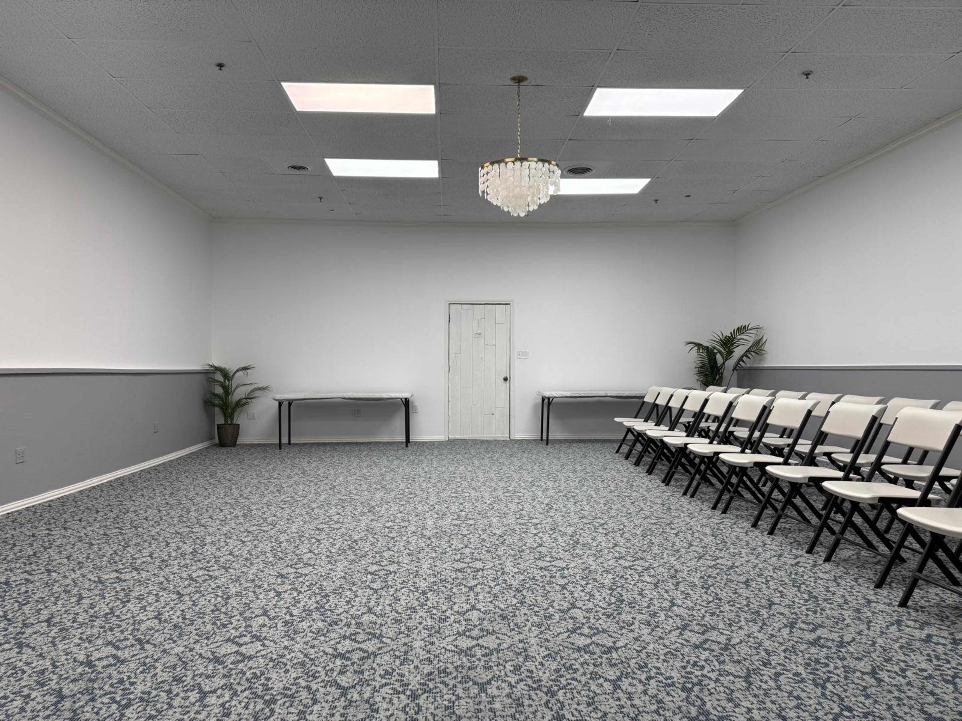 The image shows a spacious, empty meeting room with chairs arranged in a semi-circle facing a door, surrounded by plain walls and a carpeted floor.