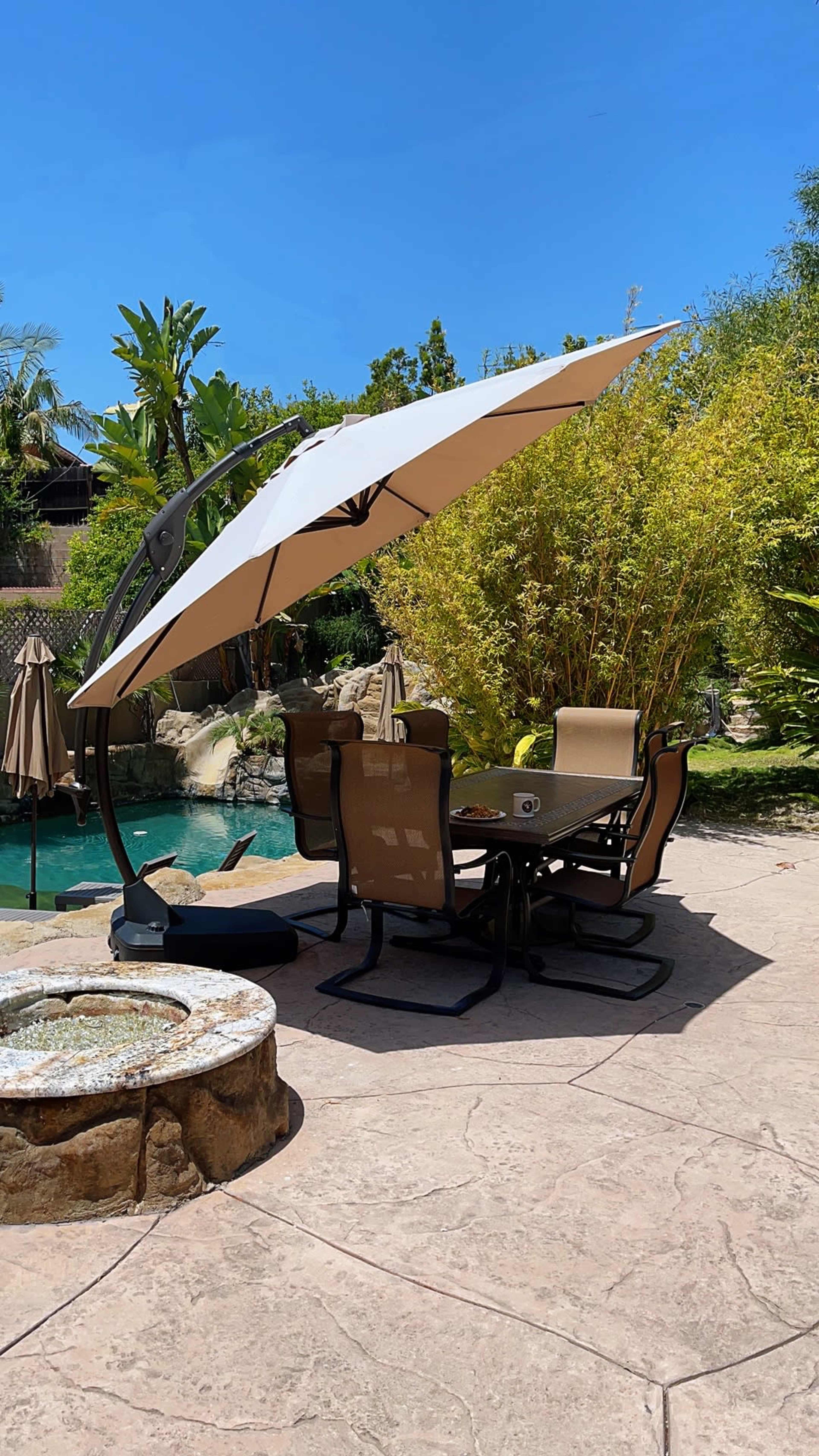 A large patio umbrella provides shade over a dining table and chairs near a pool surrounded by greenery.