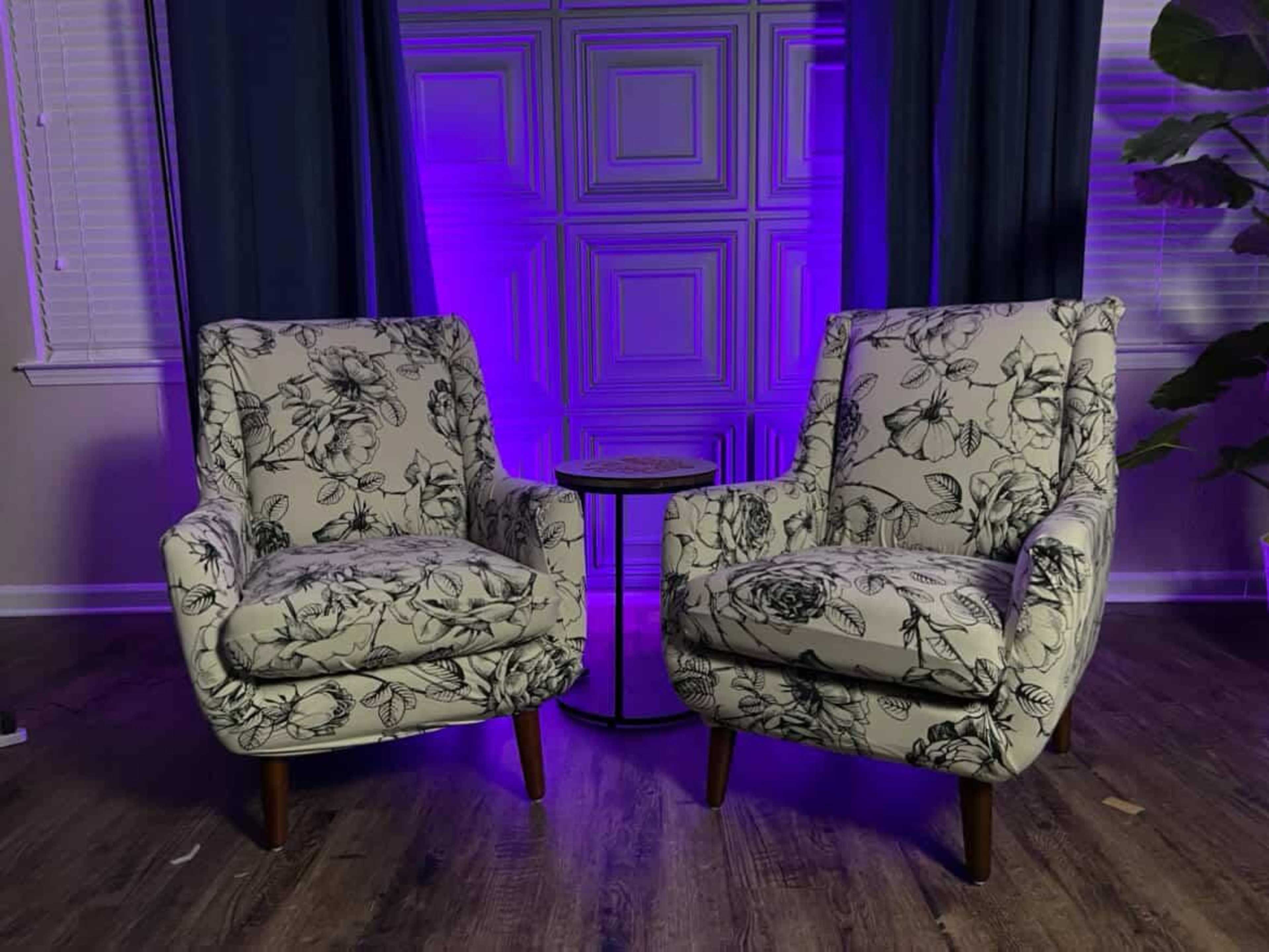 Two upholstered chairs with floral patterns are positioned facing each other across a small table in a room with purple lighting and gray walls.