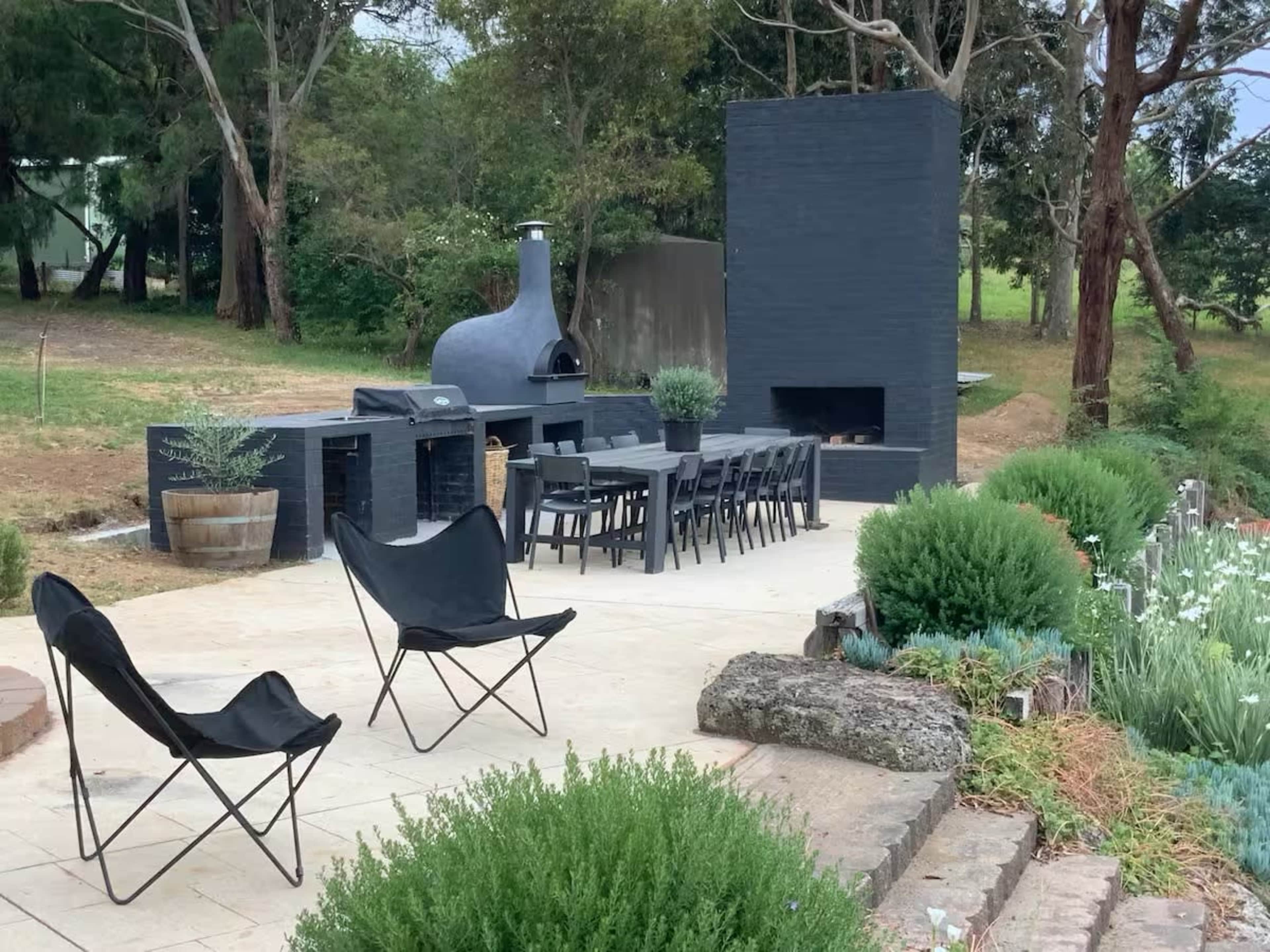 The image shows an outdoor patio area with a black brick wood-fired oven, a dining table set for several guests, and two black folding chairs surrounded by greenery and garden beds.