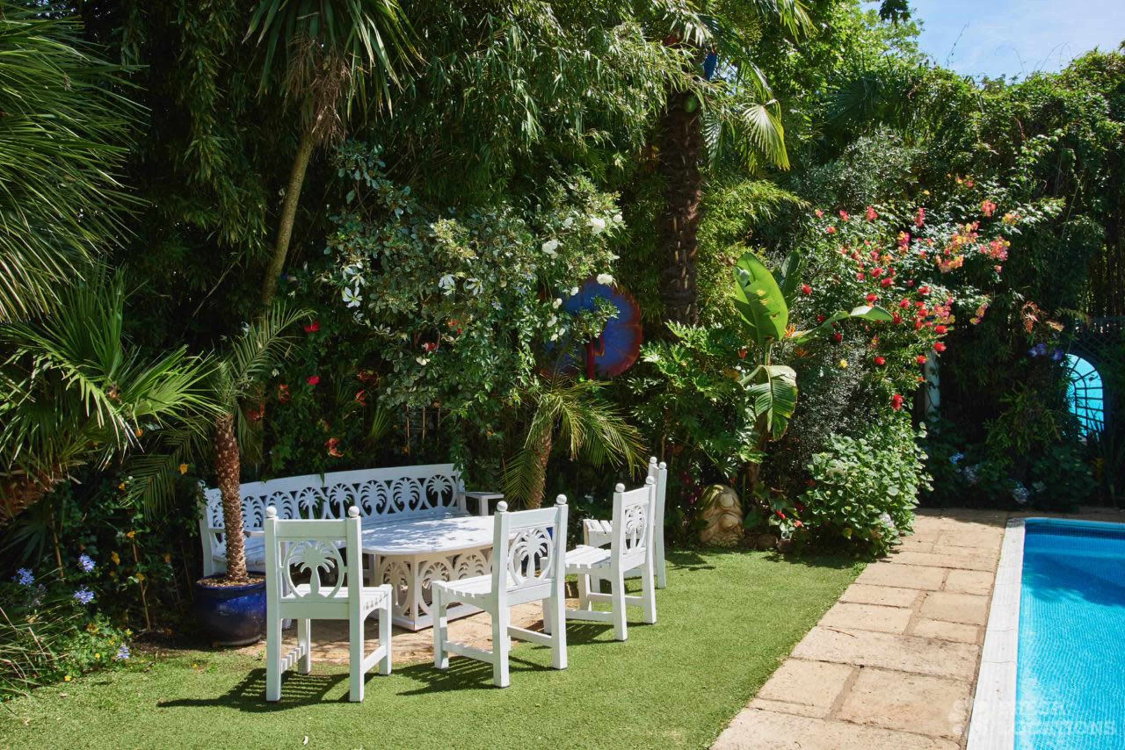 The image shows a bright patio area with a white table and chairs surrounded by lush greenery and colorful flowers next to a swimming pool.