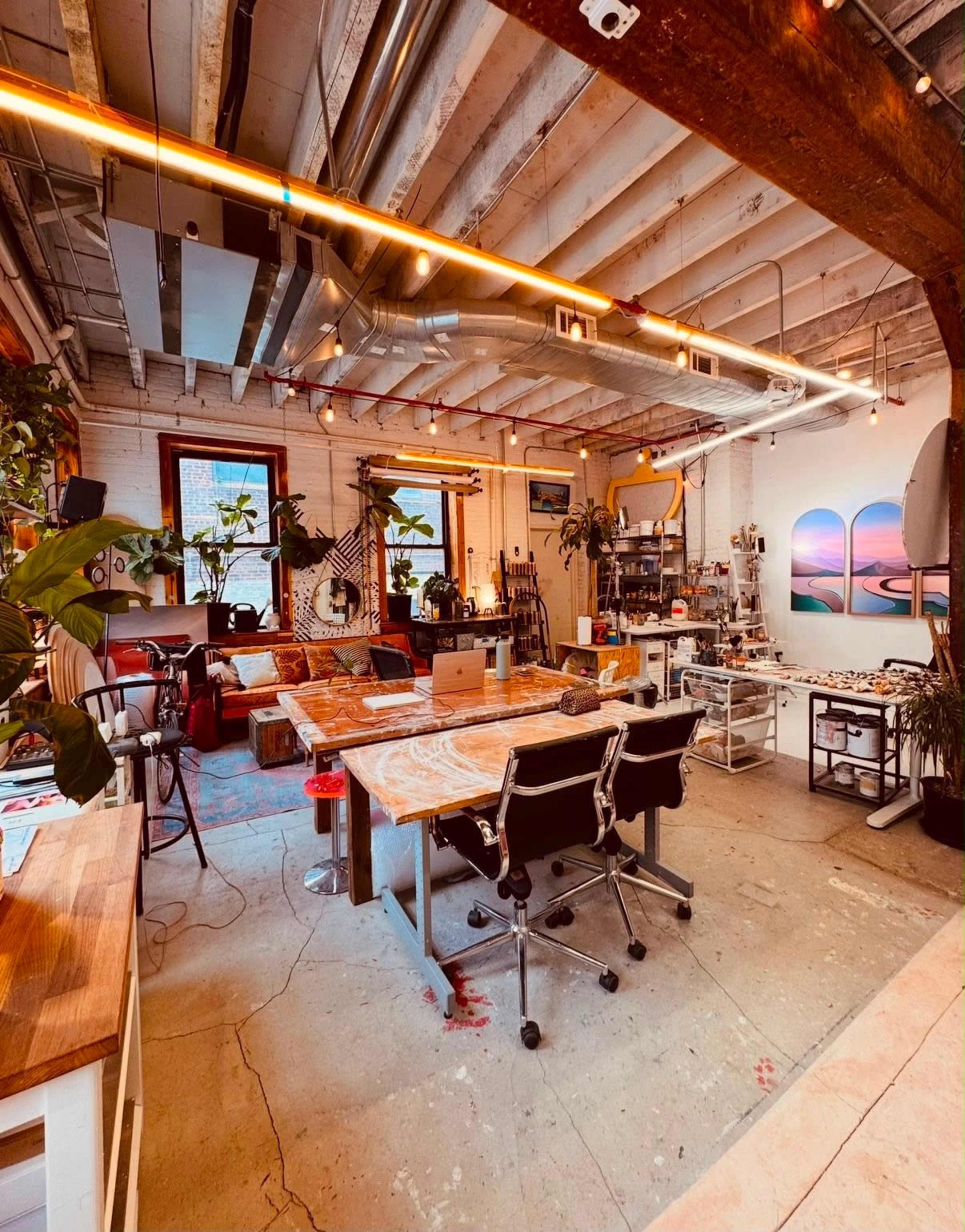 The image shows a spacious, well-lit creative studio filled with plants, featuring wooden tables, office chairs, and artwork on the walls.