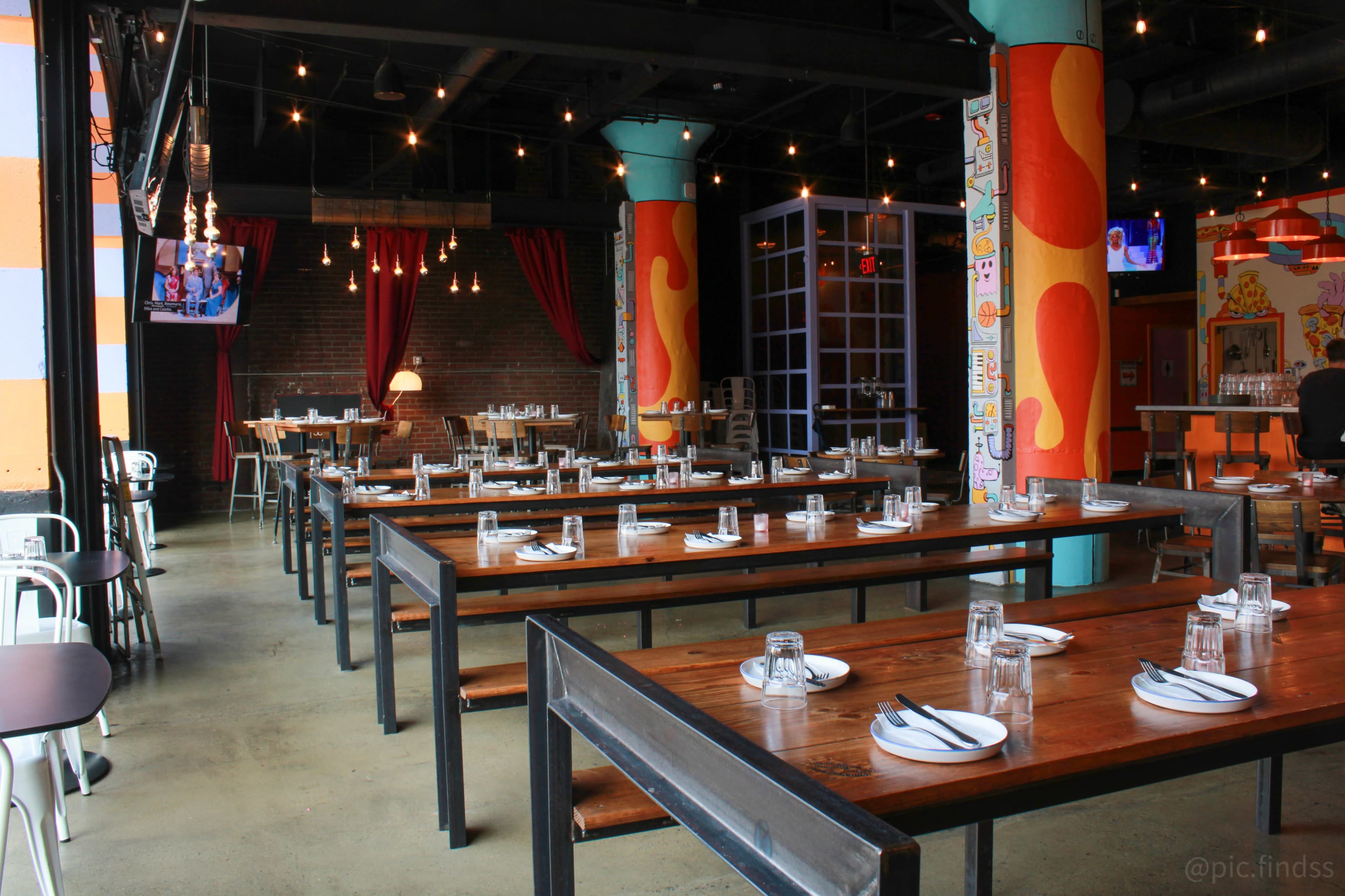 The image shows a spacious restaurant interior with wooden tables set for dining, featuring colorful pillars and soft lighting.