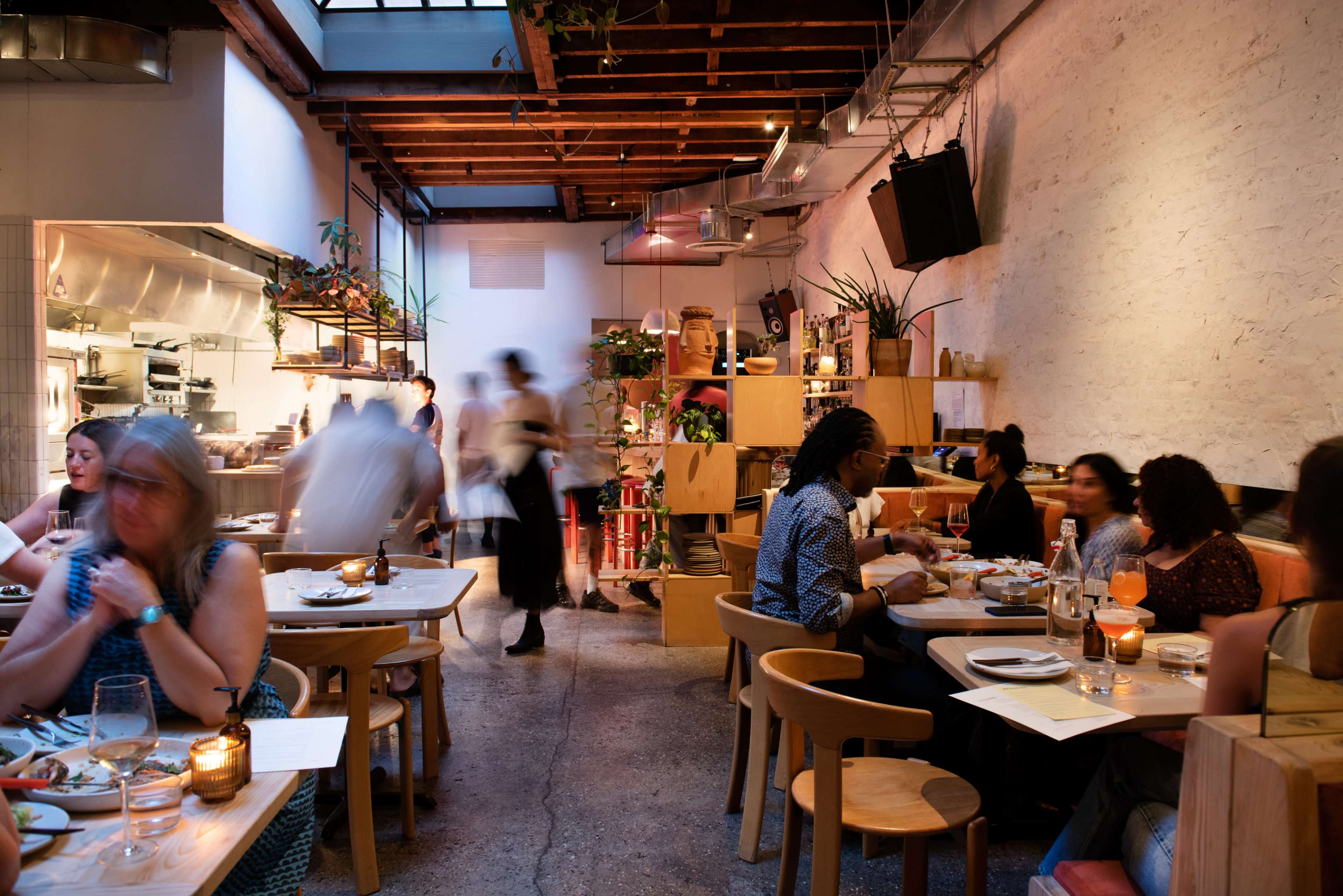 A bustling restaurant with patrons seated at wooden tables, while servers move in the background amidst a well-lit interior.