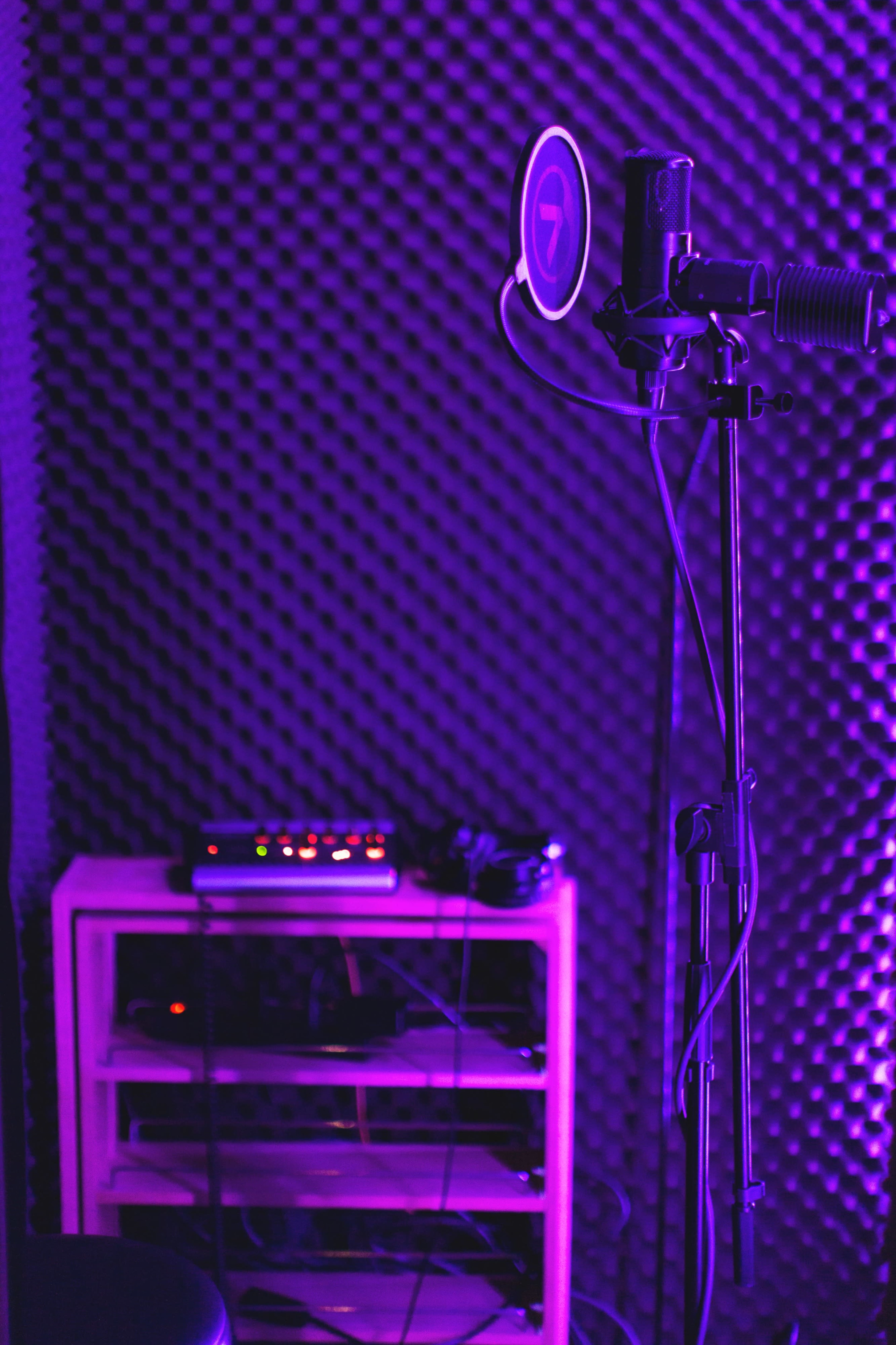 A recording studio corner with a microphone on a stand, headphones, and audio equipment on a shelf, surrounded by soundproofing foam panels.